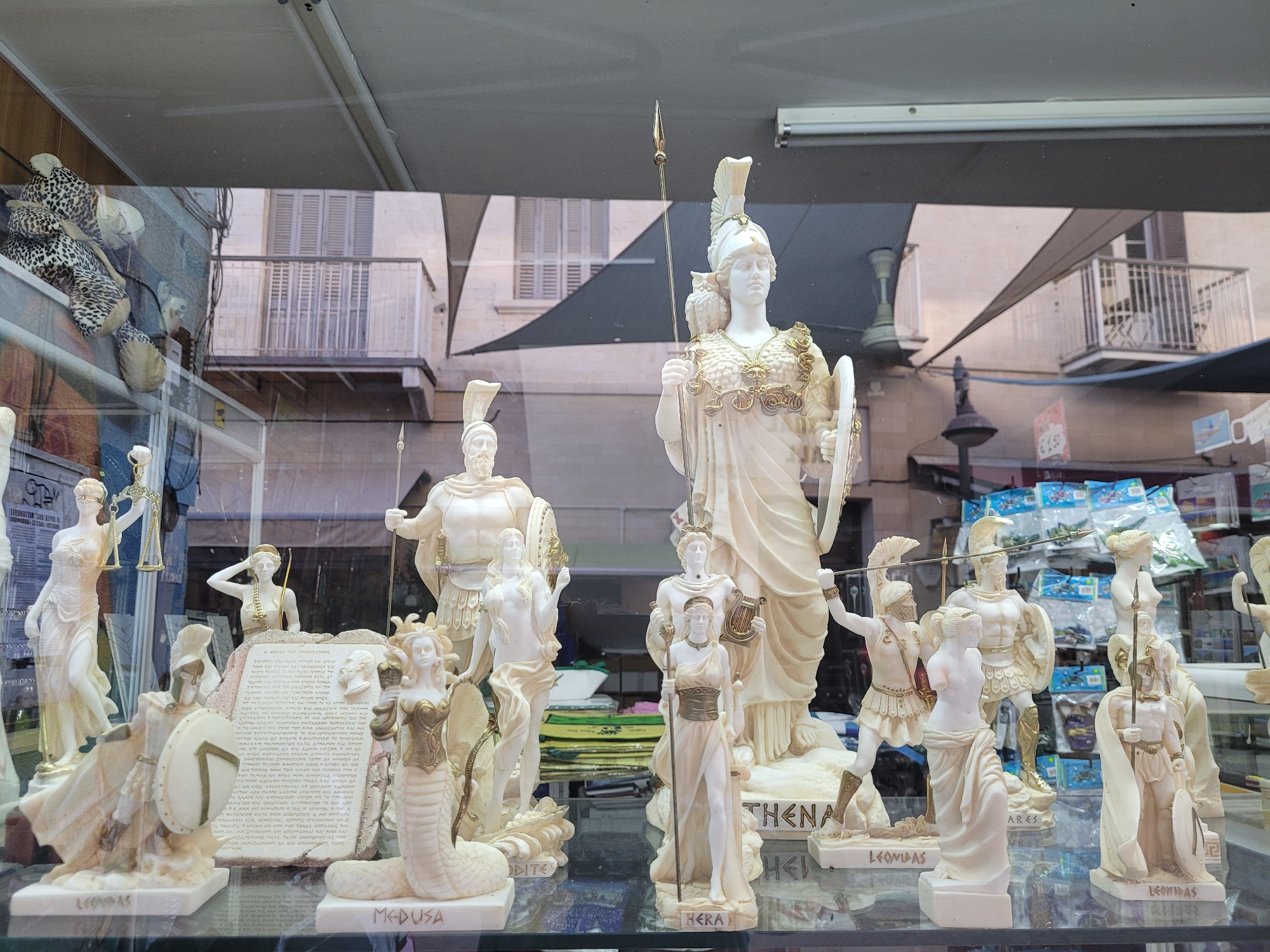 The image shows a shop window displaying a collection of white marble statues, likely of mythological figures. The statues are detailed and some are partially gilded, with elements such as armor, weapons, shields, and headdresses. In the foreground, several statues are clearly identifiable with labels on their bases, including "MEDUSA", "LEONIDAS", and "HERA". Other statues depict figures that appear to be goddesses and warriors. In the background, through the window, buildings with balconies and an awning are visible, suggesting an urban streetscape. There are also some plush toys with a leopard print pattern hanging on the left side of the window. The overall impression is of a souvenir shop or a shop specializing in decorative items, possibly located in a tourist area given the subject matter of the statues. The city of Limassol, Cyprus, is a port city with a rich history, and it is plausible that such a shop would exist there, catering to tourists interested in Greek mythology and historical figures.