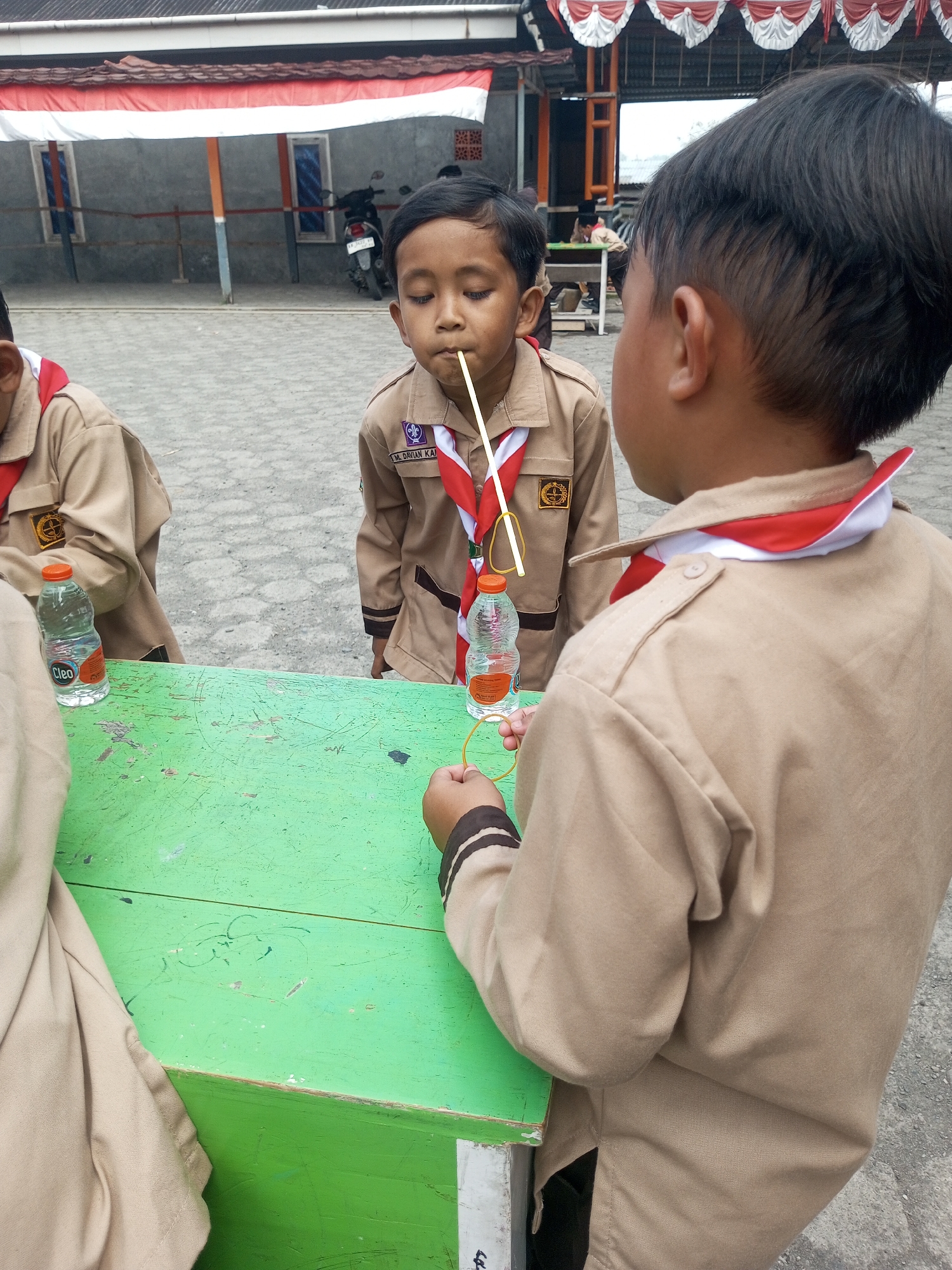 In an outdoor scene in Kalibeber, Indonesia, two young boys in tan scout uniforms with red and white neckerchiefs are engaged in an activity at a green, weathered table. The boy on the left is holding a straw in his mouth, with a rubber band looped around it and a bottle of Cleo water. The boy on the right, facing away from the camera, is looking towards the boy with the straw and appears to be holding a rubber band. The background shows a paved area, a building with a red and white banner, and a motorcycle parked nearby.