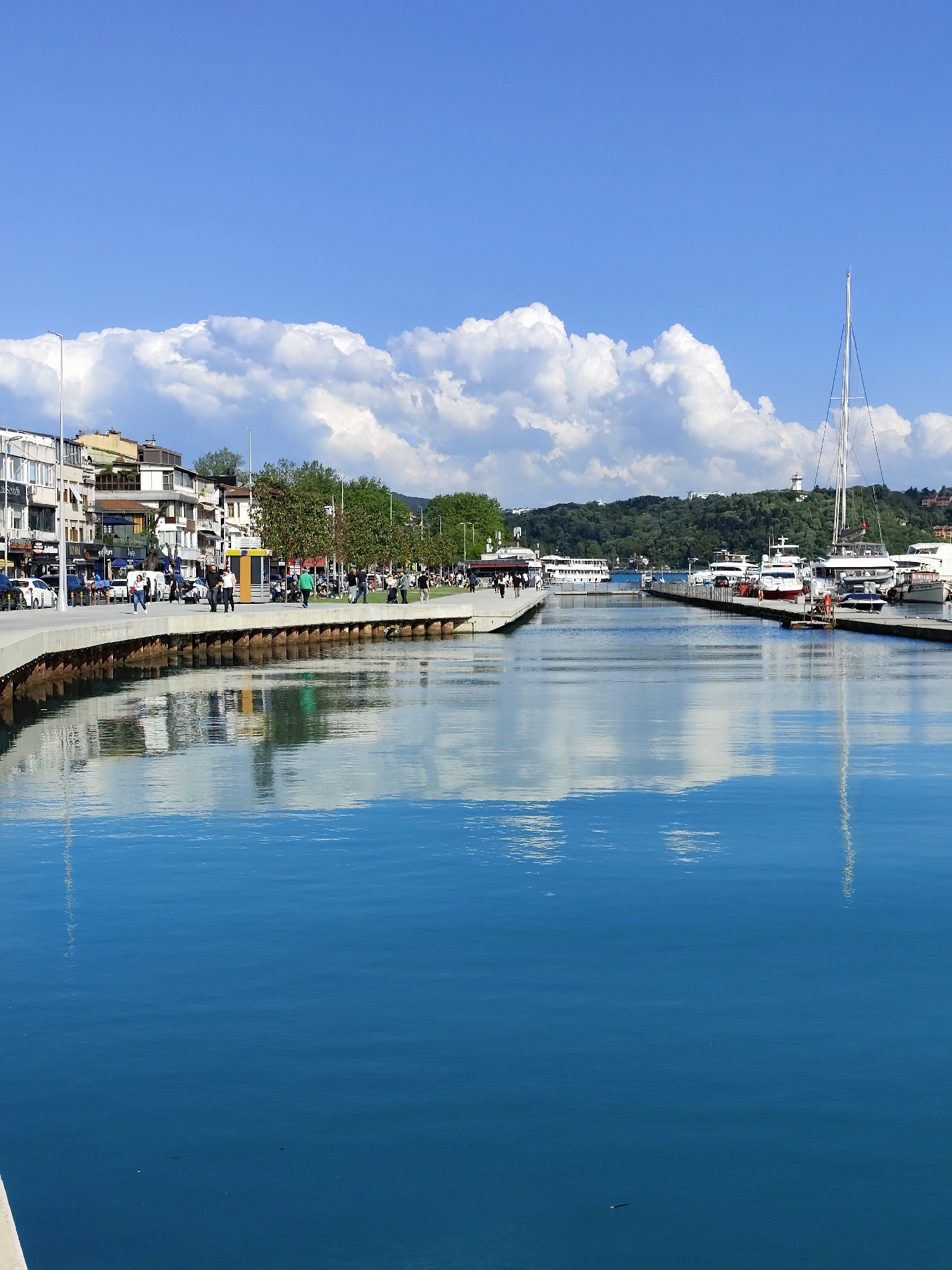 The image depicts a harbor scene with calm blue water reflecting the sky and clouds. A waterfront promenade runs along the left side, lined with buildings, trees, and streetlights. People are seen strolling along the promenade. Several yachts and boats are docked at a pier that extends into the water. In the background, a tree-covered hill can be seen, topped with a white building that may be a lighthouse. The location appears to be a coastal city.