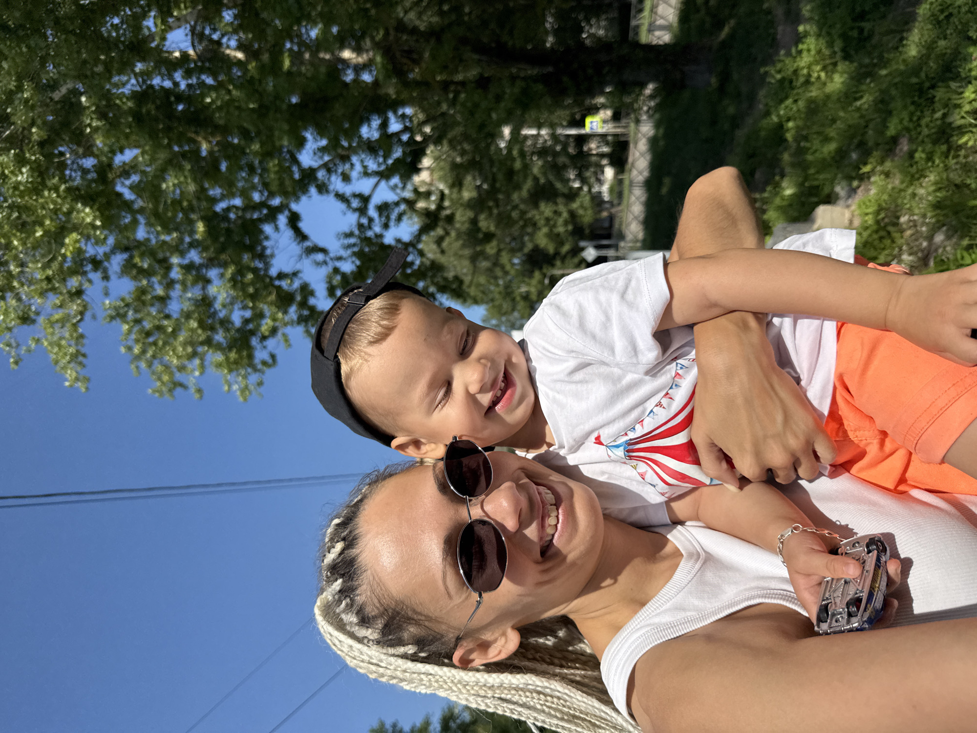 In an outdoor, sunny setting, a woman with blonde braided hair and sunglasses smiles as she holds a young child. The child, wearing a black baseball cap, a white t-shirt with a circus tent design, and orange shorts, is held closely by the woman and appears to be smiling with closed eyes. The woman is wearing a white tank top and is holding a small toy car in her left hand. In the background, there are trees, a blue sky with overhead wires, and a hint of urban infrastructure such as a street and buildings. The scene suggests a candid, joyful moment between the two individuals in a public outdoor space.