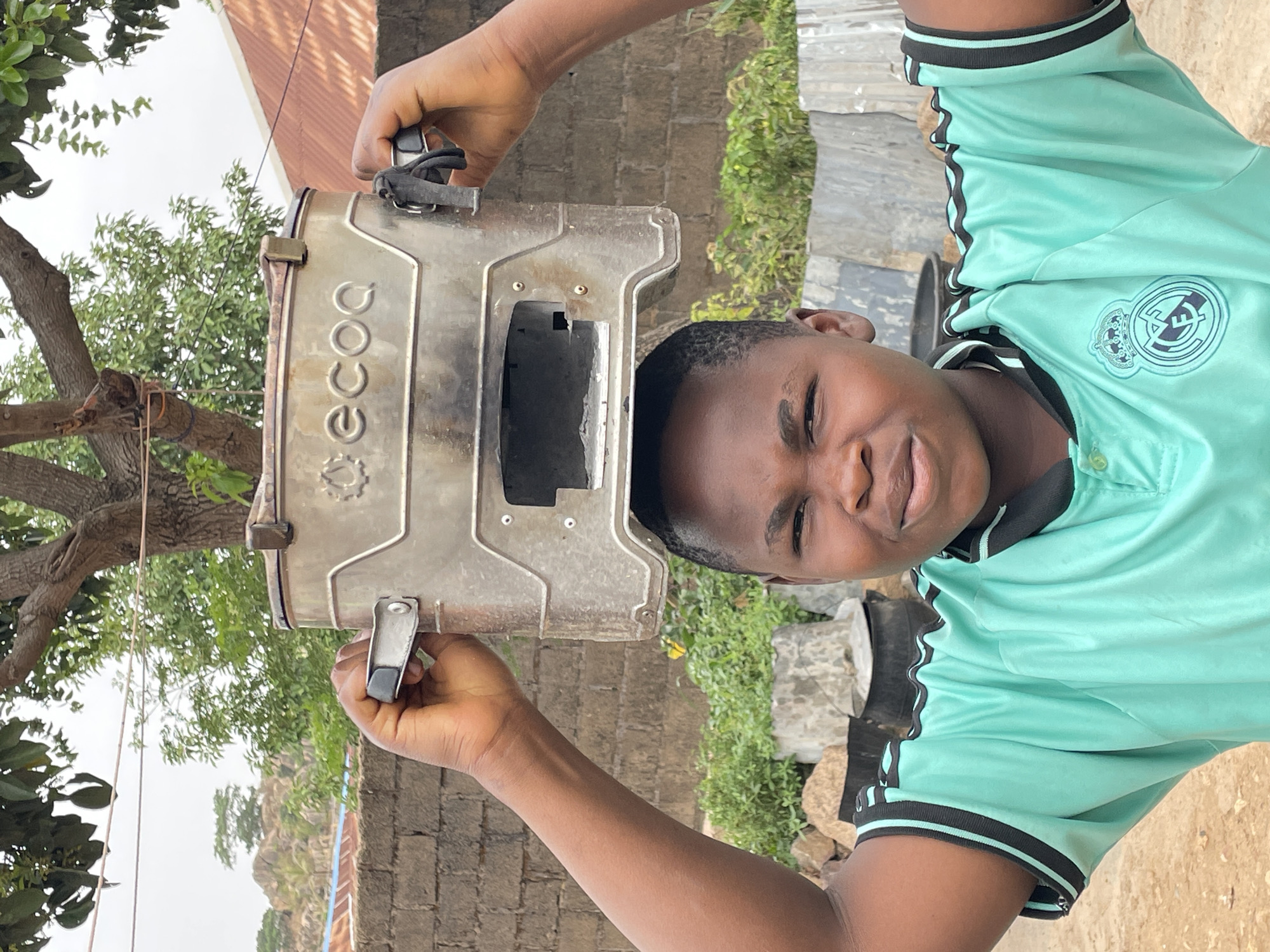 A young man is holding up a silver-colored ECOG gas stove with two latch closures. The stove has a rectangular opening in the front. He is wearing a teal-colored, short-sleeved polo shirt with black trim and the Real Madrid emblem on the chest. The background includes a brick wall, trees, vegetation, and what appears to be a rocky outcrop in the distance. The picture was taken outdoors in Jos, Nigeria.