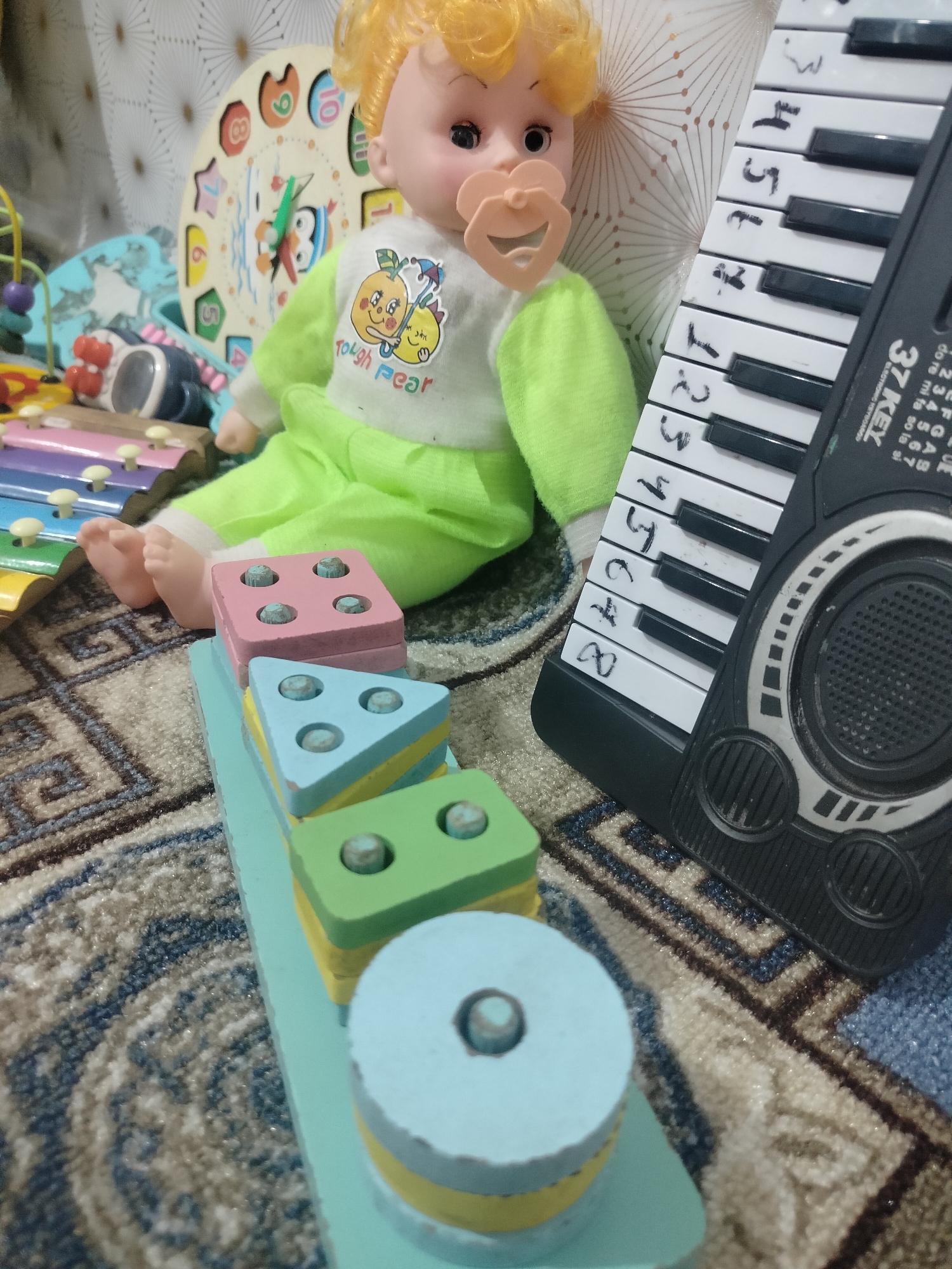 The image depicts a collection of children's toys arranged on a patterned rug. In the center is a doll with blonde hair, wearing a light green outfit with a graphic of two peaches and the text "tough pear." The doll has a pacifier in its mouth. To the left of the doll, a colorful xylophone is partially visible. To the right of the doll is a black electronic keyboard with white keys, some of which have numbers or symbols written on them in black marker. In the foreground, a wooden shape-sorting toy is stacked with colorful blocks: a pink square, a blue triangle, and a green square, each with pegs. A light blue circular block rests at the bottom. The background includes a circular clock-like toy with numbers and cartoon characters. The overall scene is of a child's play area, suggesting a domestic indoor setting. There is no specific information in the image that would indicate its location in the city of Cholpon, Kyrgyzstan.