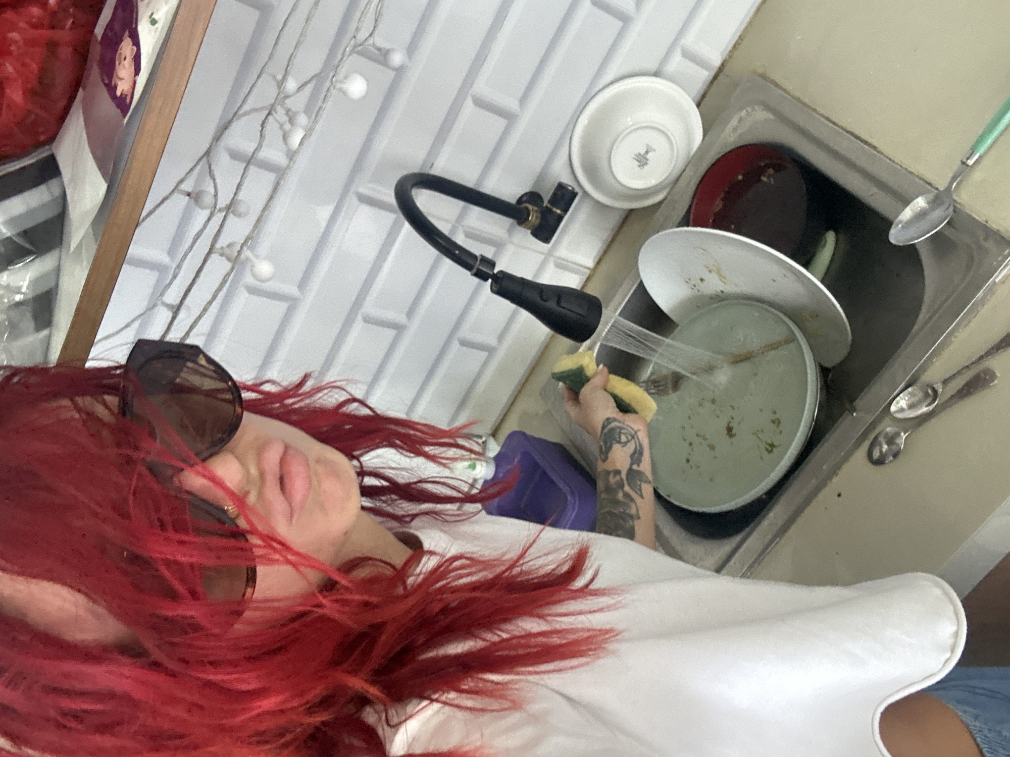 The image, taken in what is noted as North Kuta, Indonesia, shows a close-up, first-person perspective of a person in a kitchen setting, engaged in washing dishes.
The central subject is a person, likely a woman, with striking, long, bright red hair that cascades around her face and shoulders. She wears dark, large-framed sunglasses that obscure most of her eyes, and her lips appear lightly colored. She is dressed in a plain white top and what appears to be blue denim shorts or a skirt, partially visible at the bottom right. Her left arm, prominently featured, displays extensive dark tattoos, and her hand is holding a yellow and green cleaning sponge.
The activity taking place is dishwashing. The person's hand with the sponge is positioned directly over a stainless steel kitchen sink that is filled with dirty dishes. Visible items in the sink include several plates with food residue, a dark red bowl, and a light-colored bowl flipped upside down. Water appears to be actively running onto one of the plates from the black gooseneck kitchen faucet. A spoon with a light green handle rests on the edge of the sink, and other spoons are visible on the counter beside it.
Notable details include the white, rectangular-tiled backsplash behind the sink, giving a clean and modern appearance to the wall. Decorative string lights with small white spherical bulbs are draped in the background. A wooden frame or shelf edge is also visible on the left side of the frame. A purple plastic container is placed near the sink. On the underside of one of the white bowls, faint text, possibly "WEDGWOOD," is visible. The scene captures a moment of domestic chore.