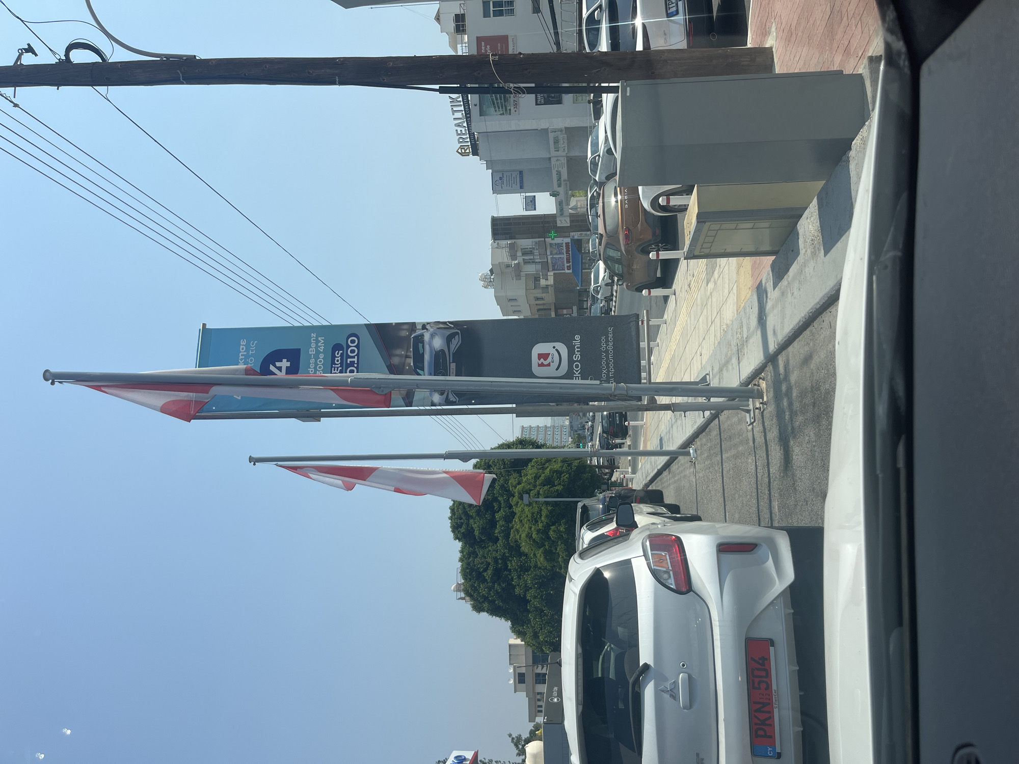 The image shows a street scene in Germasogeia Municipality, Cyprus. A white Mitsubishi Mirage is parked in the foreground with a license plate reading "PKN 504 CY". Behind it, several other cars are lined up on the road, suggesting traffic congestion. A large banner advertising "EKO Smile" and a Mercedes-Benz 300e 4M is visible on the right side of the street. The banner also displays pricing information. Several flags, including what appear to be Cypriot flags, are flying from poles. Buildings, trees, and a wooden utility pole are present in the background. The sky is clear and blue, indicating a sunny day. The scene appears to be a typical urban environment with commercial establishments.
