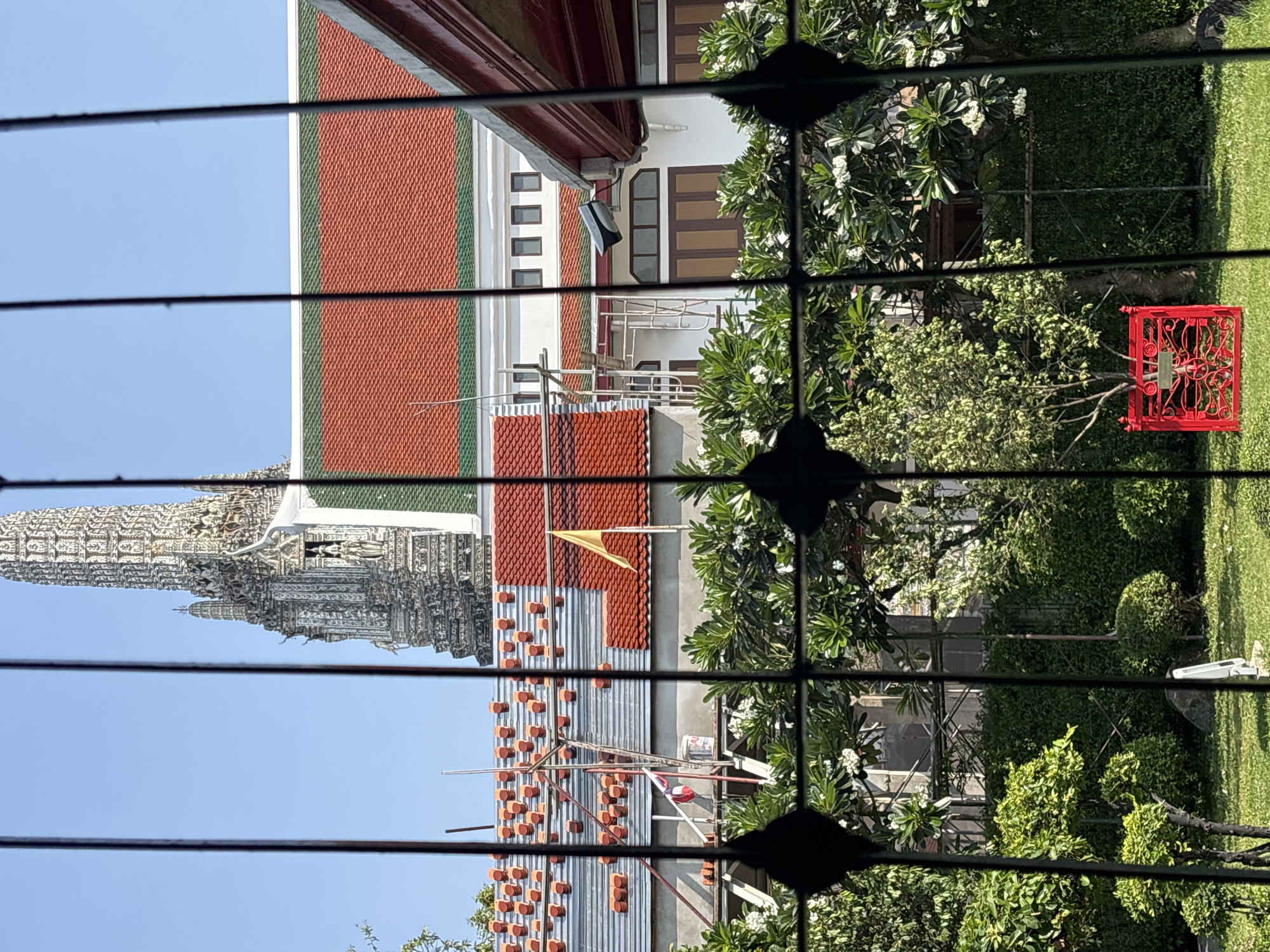 View through a window with a grid pattern reveals Wat Arun (Temple of Dawn) in Bangkok, Thailand. The temple's distinctive white prang is visible against a clear blue sky. Nearby buildings with red-tiled roofs and white walls are also in view. Lush green foliage and a red ornamental structure stand out in the foreground. Construction activity is observed, including scaffolding and new roof tiles.