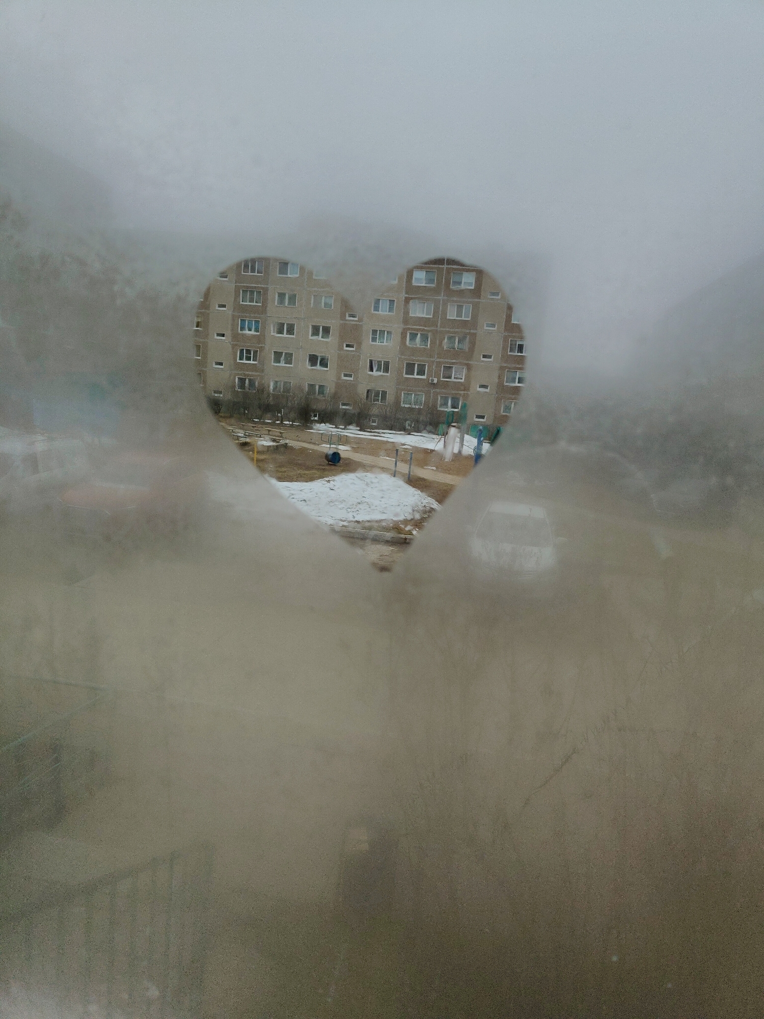 A view through a heart-shaped cutout in what appears to be a fogged or frosted window. Beyond the cutout is a residential building with multiple floors and windows. There is some snow on the ground near the building, and playground equipment is visible. A car is also visible through the heart. The surrounding area is obscured by the window frost.