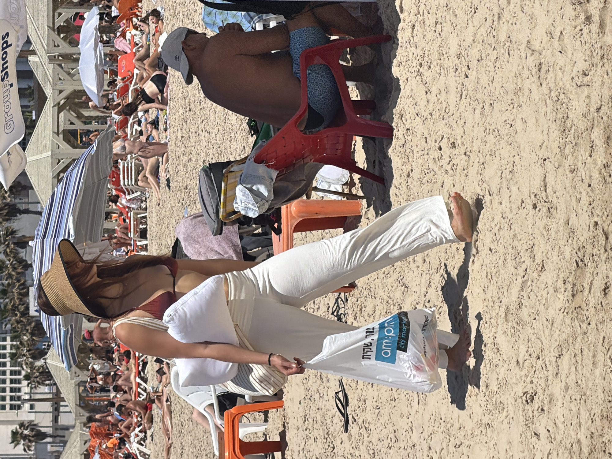 The image depicts a crowded beach scene in Tel Aviv, Israel. Numerous people are sunbathing and relaxing under striped umbrellas and canopies, seated in orange and white beach chairs.
In the foreground, a woman wearing a straw hat, a brown bikini top, and white pants is walking. She is carrying a white plastic bag with Hebrew lettering. A pair of black flip-flops lies on the sand near her feet.
Behind her, a shirtless man wearing a hat and patterned blue shorts sits on a red plastic chair. His back is toward the camera.
The beach is sandy, with a light brown hue. In the background, a building is visible. The day appears sunny and bright.