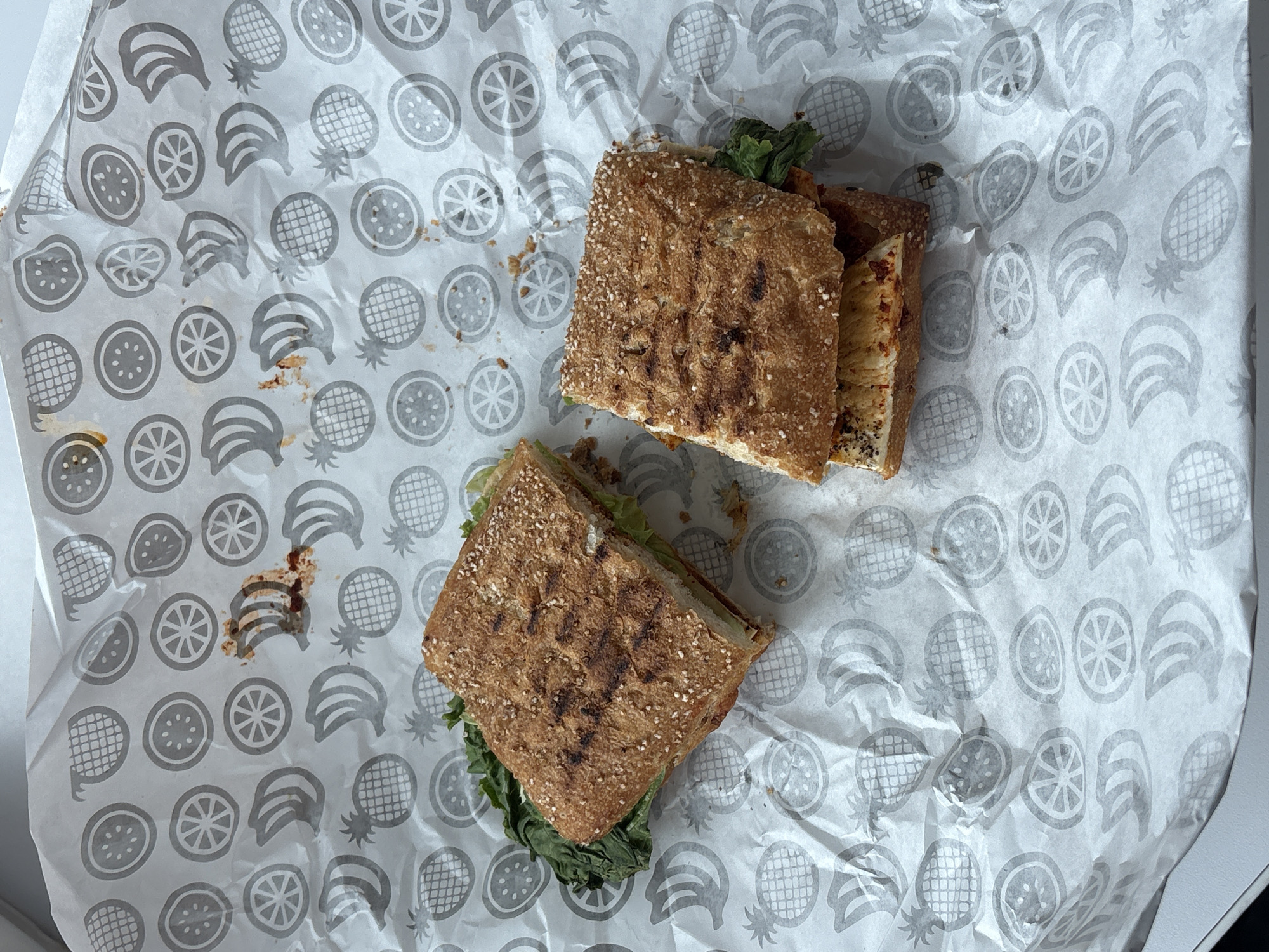 The image shows two halves of a panini sandwich on a sheet of white patterned paper. The paper has a repeating pattern of grey stylized fruits, including citrus slices, bananas, and pineapples. The sandwich halves appear to have been pressed, with grill marks visible on the bread. One half has lettuce and possibly other fillings, while the other shows filling with a yellow and red hue. There is some sauce spillage on the paper near the lower sandwich half. The context of Puebla, Mexico, is not evident from the image itself.
