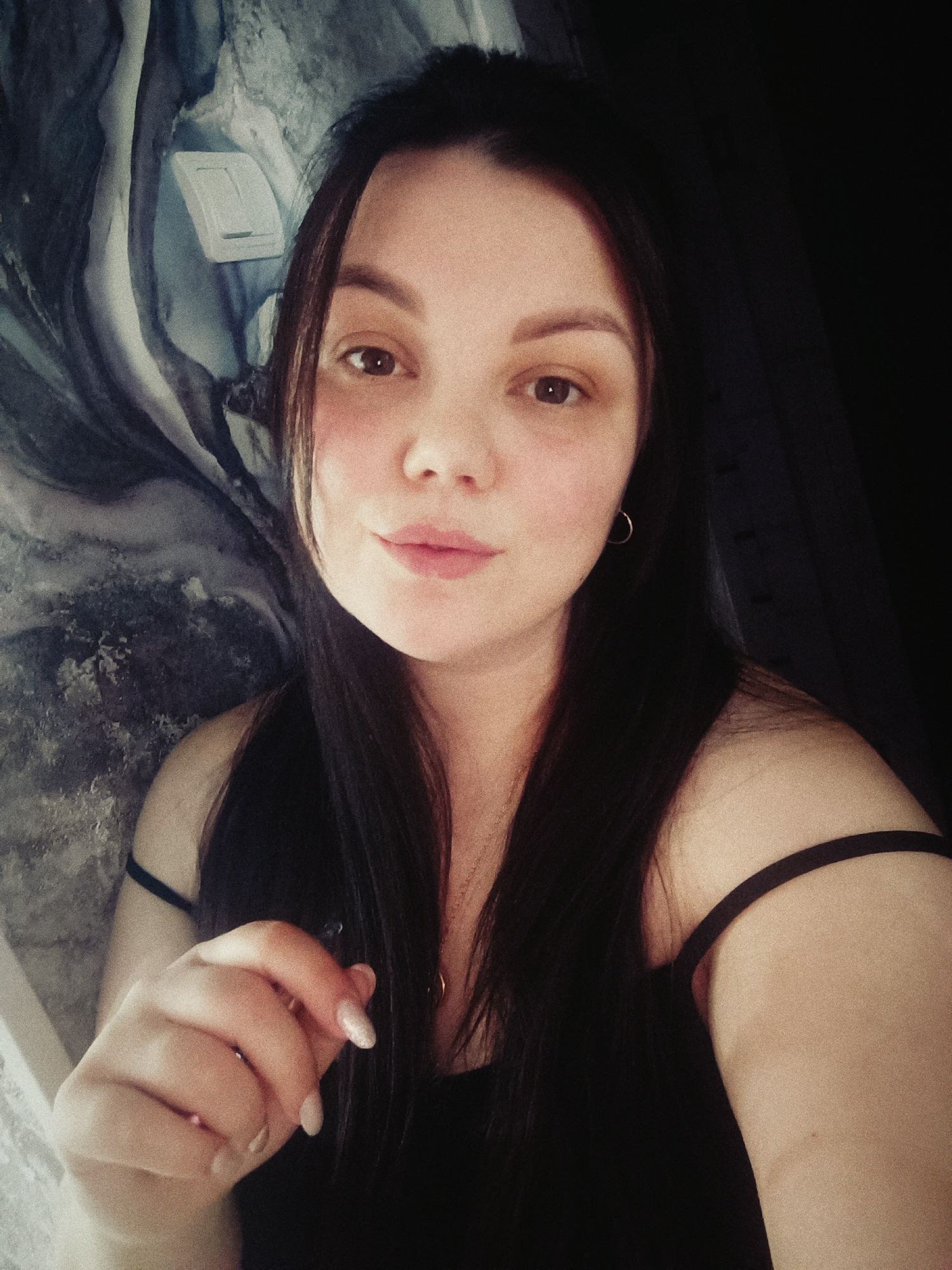A medium close-up shot of a woman with dark hair and fair skin. She is looking directly at the camera with a neutral expression. Her lips are slightly parted. She is wearing a black tank top. Her left hand is in the foreground, partially obscuring her torso. Her nails are manicured with light polish.
Behind her, there is a textured wall or curtain with a marbled pattern in shades of grey and blue, along with what looks like a light switch. To her right, there is a dark, undefined area, possibly a wall or heavy curtain, that suggests a dimly lit room.
There is no information indicating the city of Chaikovsky, Russia.