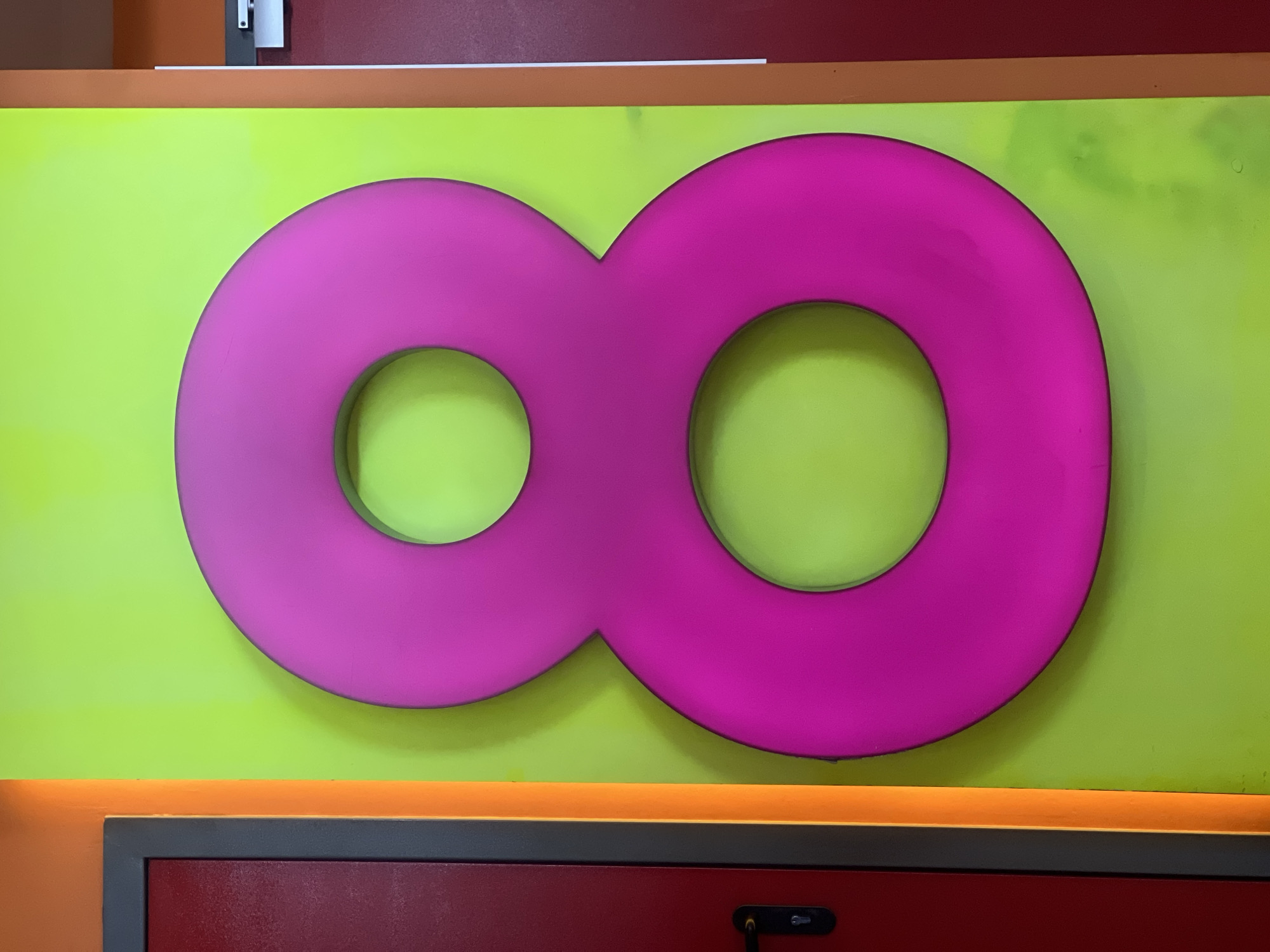 A pink infinity symbol is mounted on a bright green wall with visible smudges and discoloration. The wall is bordered by an orange horizontal stripe above a red panel with a dark gray trim. A black object, possibly a handle, is visible on the red panel. Above the green wall, there's a glimpse of a reddish-purple surface and a white object with a gray surround. There are no people visible in the image. The location is Krasnoyarsk, Russia.