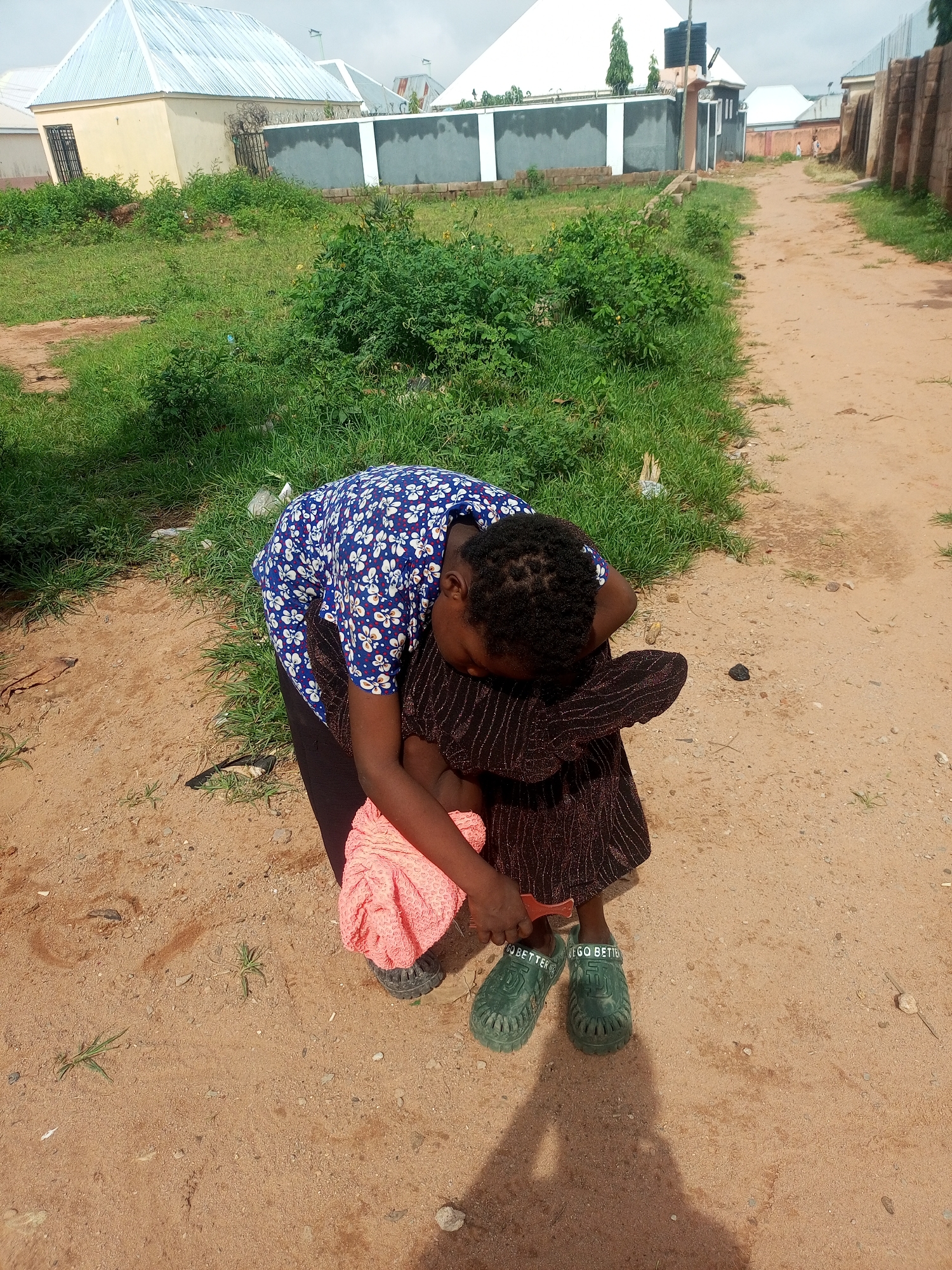 A young person is hunched over on a dirt road in Tempure, Nigeria. They are wearing a blue and white floral shirt, a dark, possibly sequined, skirt, and green slip-on shoes with the inscription "GO BETTER" visible on them. The person appears to be holding a pink garment. The ground is a mixture of sand and small pebbles, with scattered green grass and weeds. In the background, there are buildings with corrugated metal roofs, a wall, and a dense patch of green foliage. The sky is overcast.