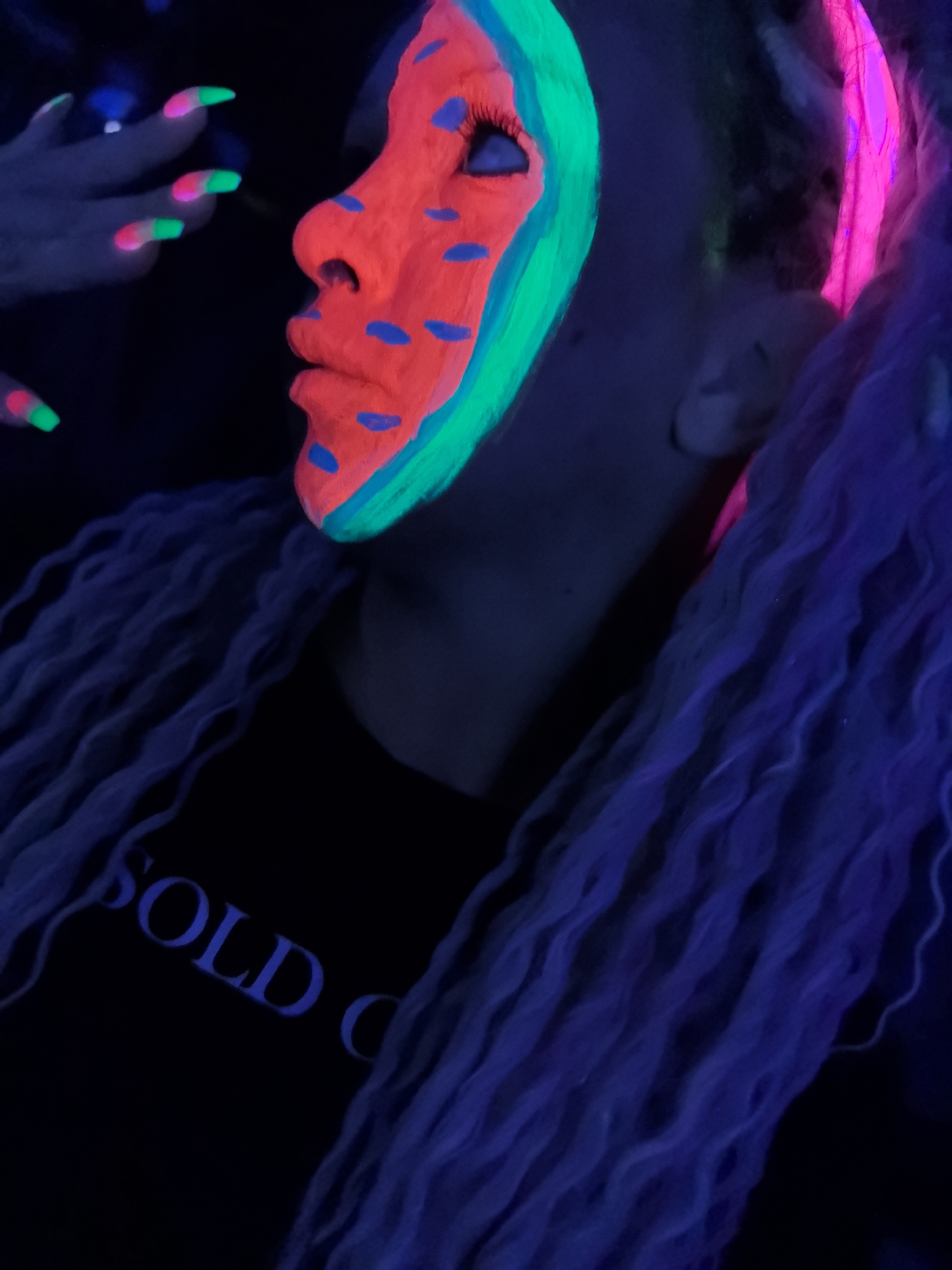 The image shows a person in a dark environment illuminated by ultraviolet light. The person's face is painted to resemble a watermelon, with a red interior and blue seed-like spots. A green line is used as a rind along the edge of the face. The person has long, wavy, light-colored hair that is also illuminated by the UV light. The person's nails on one visible hand are painted with a gradient effect, with bright green tips fading into a pinkish hue. The person is wearing a dark t-shirt with the word "SOLD" visible in blue letters. There is no discernible information to place this scene in the city of Noviy, Russia.