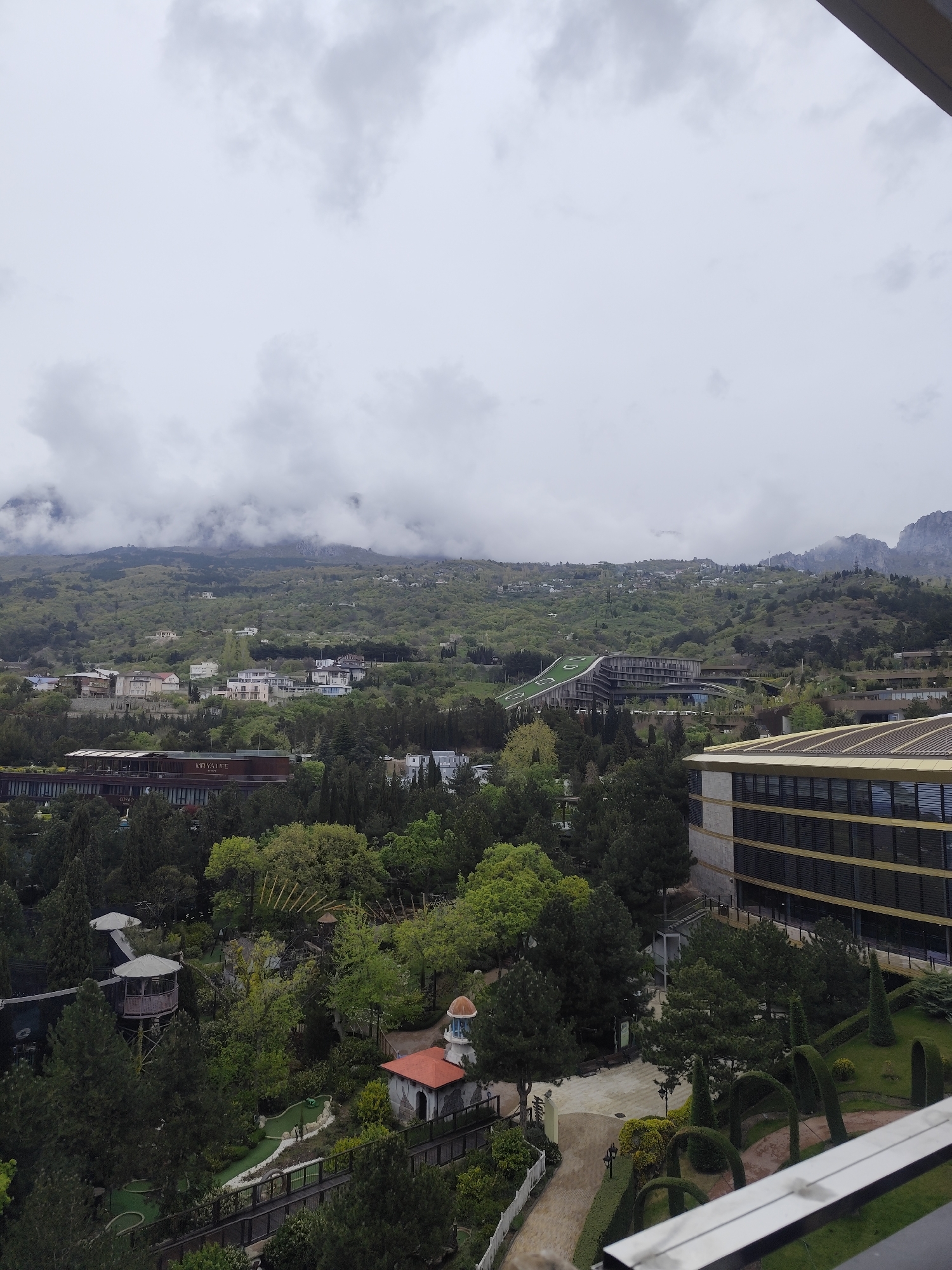 An expansive, high-angle outdoor shot captures a vast landscape under an overcast sky, likely in the mountainous region of Симеизский поселковый совет, Ukraine. The sky is filled with light grey clouds, some hanging low and partially obscuring the rugged, green-covered mountains in the distant background.
The mid-ground is dominated by lush green hillsides densely covered with trees and scattered residential buildings. A prominent modern building with a distinctive green, sloped roof is visible on a hillside, blending somewhat with the natural contours. Closer to the foreground, another large, multi-story building features a contemporary design with dark-tinted windows and golden-brown horizontal accents. The text "MRIYA LIFE" and "CASINO" are visible on this building.
The immediate foreground reveals a meticulously landscaped area featuring numerous mature evergreen trees, manicured hedges, and paved pathways. There are several decorative architectural elements, including a small domed building with a terracotta-colored roof and a gazebo-like structure on stilts. A mini-golf course with artificial turf and small obstacles winds through the green space, crossed by a wooden bridge or walkway. The overall impression is one of a resort complex nestled within a natural, mountainous environment. There are no people visible in the image. The cloudy weather suggests a cool or temperate day.