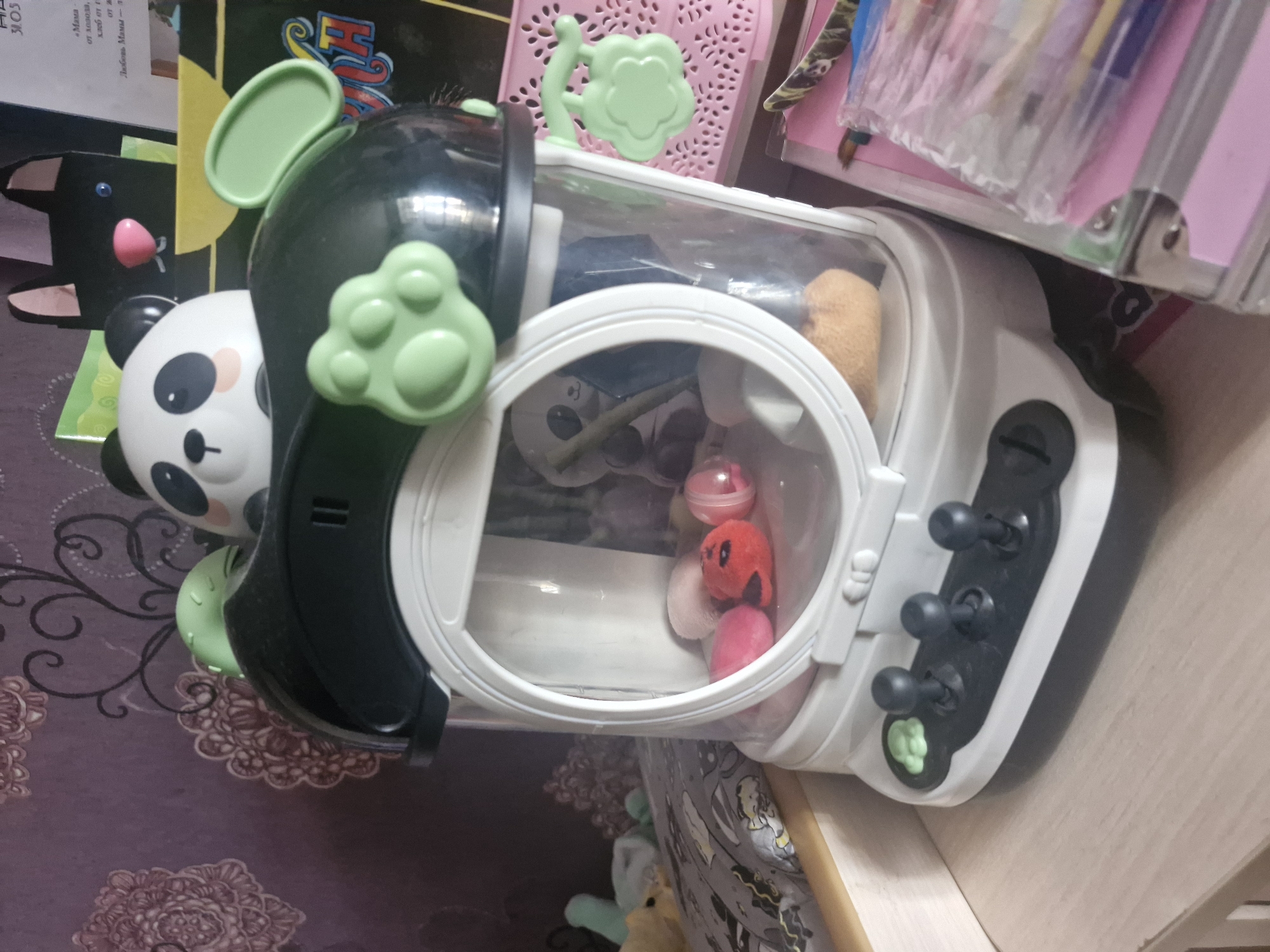 The image shows a toy claw machine in the shape of a panda. The machine is white and black, with green accents. The claw is visible inside the machine, along with several small stuffed animals. The machine is sitting on a surface, possibly a table or shelf, with other items visible in the background. The location and city are not identifiable from the image.