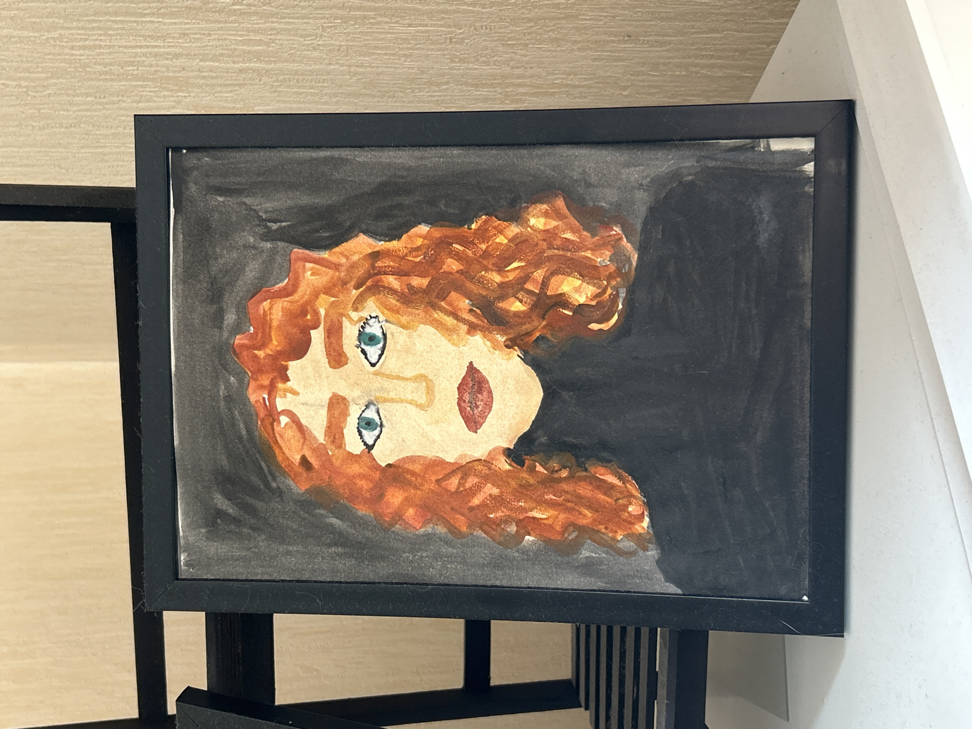 The image displays a framed watercolor painting of a woman with bright orange, curly hair. She has light skin, green eyes, and red lips, and is wearing a black turtleneck. The background of the painting is dark gray, giving it a somewhat somber mood. The painting is enclosed in a thin, black frame. The framed artwork is positioned on a white surface, possibly a shelf or table, with a textured, light-colored wall behind it. In the background, to the left of the framed painting, a dark metal structure with horizontal slats is partially visible, suggesting it might be a shelving unit or part of a display. There is no discernible information or visual cues within the image to indicate its location in Moscow, Russia.