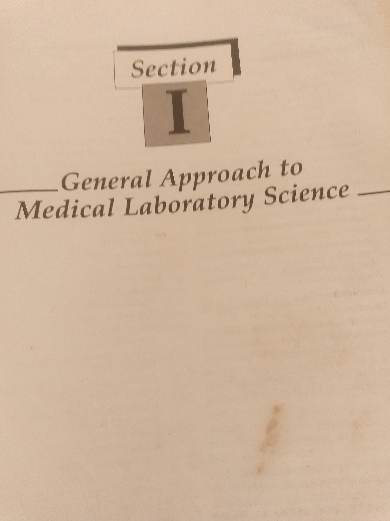 The media file is a close-up, eye-level shot of a book or document with text. The background is a light, textured paper, possibly aged, with a few faint brown spots. At the top, a banner-like graphic contains the word "Section" in a serif font. Below this, centered, is a large, gray rectangle with the Roman numeral "I" in a bold, black serif font. Beneath the Roman numeral, two lines of text are visible. The first line reads "General Approach to" in a slightly larger serif font. The second line, in a similar font and size to the first, reads "Medical Laboratory Science". A thin, horizontal line extends from the left side of "General Approach to" and a dash follows "Medical Laboratory Science". The lighting is neutral, and no specific time of day or weather conditions can be inferred. The setting appears to be indoors, likely a library or study area. There are no people or animals visible. The image focuses solely on the text and its presentation. The overall tone is educational and informative. The visible text clearly indicates the start of a new section in a publication related to medical laboratory science. The location context provided is Jalingo, Nigeria, but there are no visual cues within the image itself to support this location.