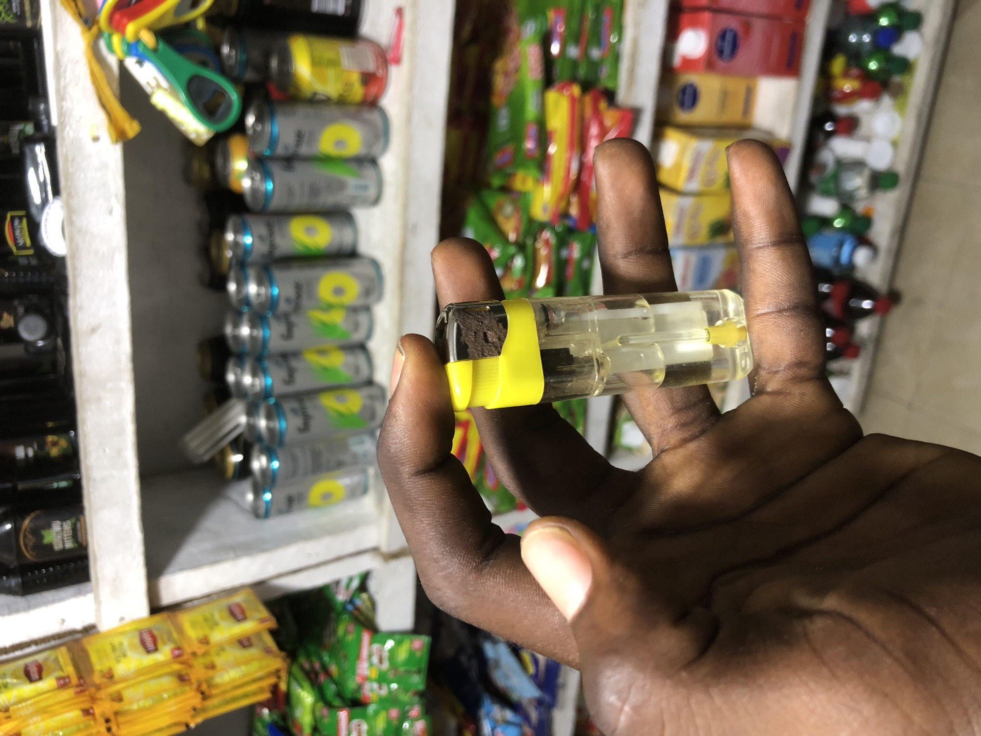 In Ado, Nigeria, a person's dark-skinned hand is holding a clear disposable lighter filled with yellow fluid. The lighter has a yellow cap and a metallic tip. The background reveals shelves stocked with various items, including packaged goods like Lipton tea sachets, canned beverages, and colorful boxes of products. The scene suggests a retail environment, possibly a small shop or market stall.