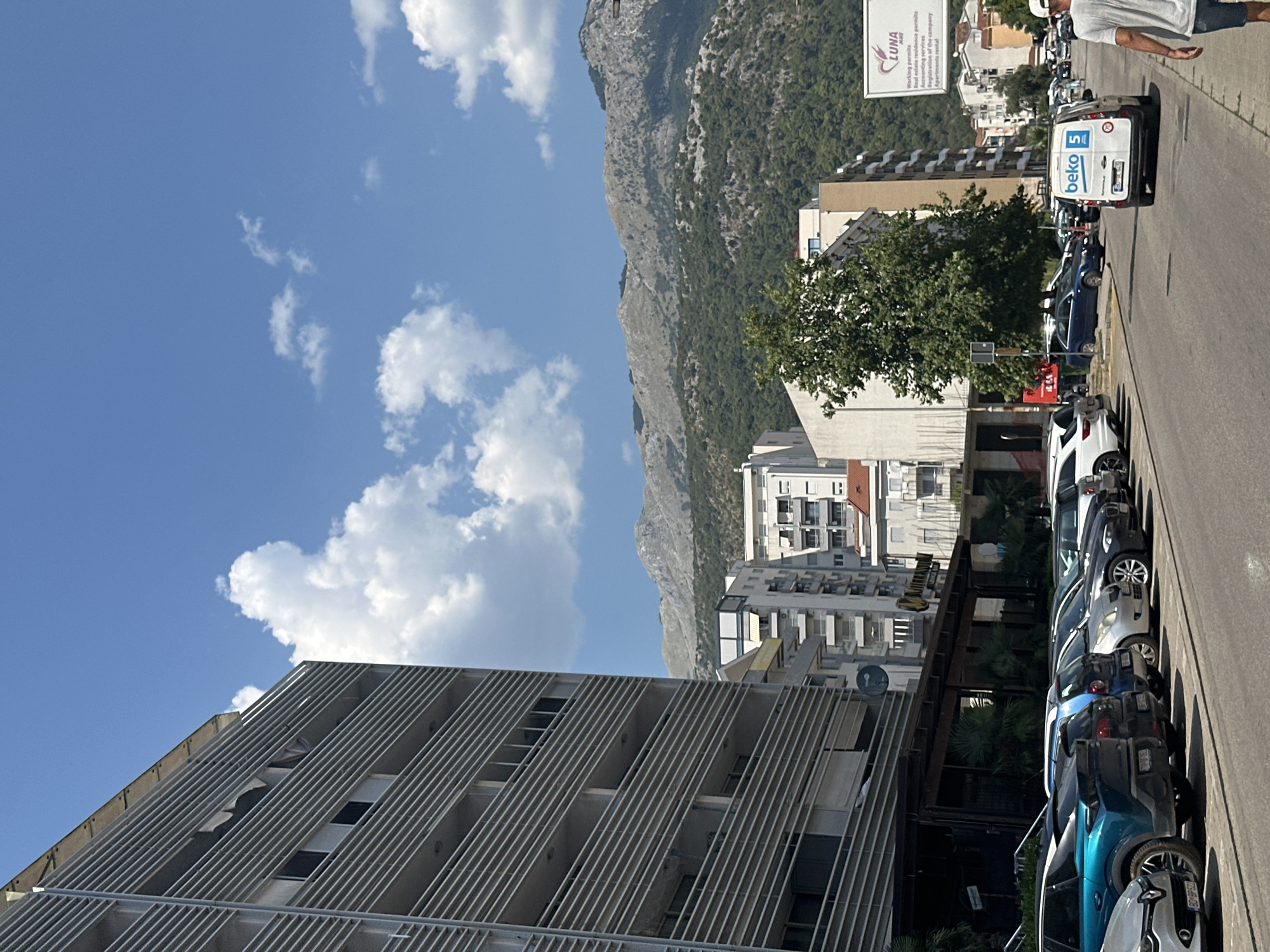 In Budva, Montenegro, along a city street lined with modern buildings and parked cars, a white van with "beko 5" branding drives down the road. On the right side of the street, a man wearing a white cap and grey t-shirt walks away from the camera. A billboard for "LUNA" is visible on the right, advertising working permits and apartment rentals. In the background, lush green trees and rocky mountains create a scenic backdrop under a clear blue sky dotted with white clouds. The scene captures a typical day in this coastal city.
