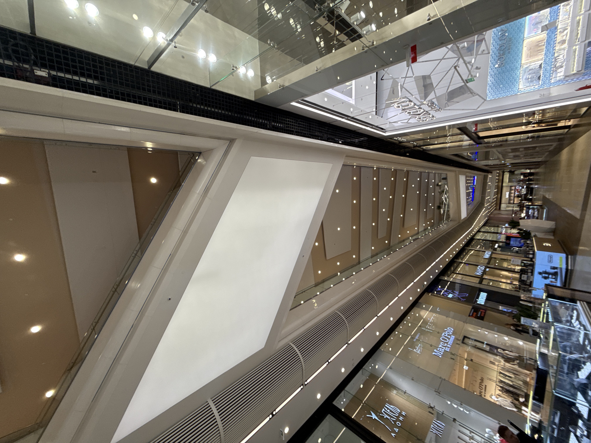 The image shows an interior view of a modern shopping mall, possibly in Moscow, Russia, with a high, multi-level ceiling featuring diffuse lighting and angular architectural elements. The perspective is wide-angle, looking upwards along a corridor lined with storefronts. On the left, the storefronts include displays with clothing racks and prominent signage for "Marc O'Polo" and "MANGO." The "Marc O'Polo" signage also indicates "Est. in Stockholm." Further down the corridor, a "SAMSUNG" sign is visible. On the right, another storefront displays the brand "Sokolov" with a blue, patterned facade and product displays. The mall's interior is characterized by glass facades, polished floors, and a grid-like ventilation system along the ceiling. The overall impression is one of a bright, spacious, and contemporary retail environment.