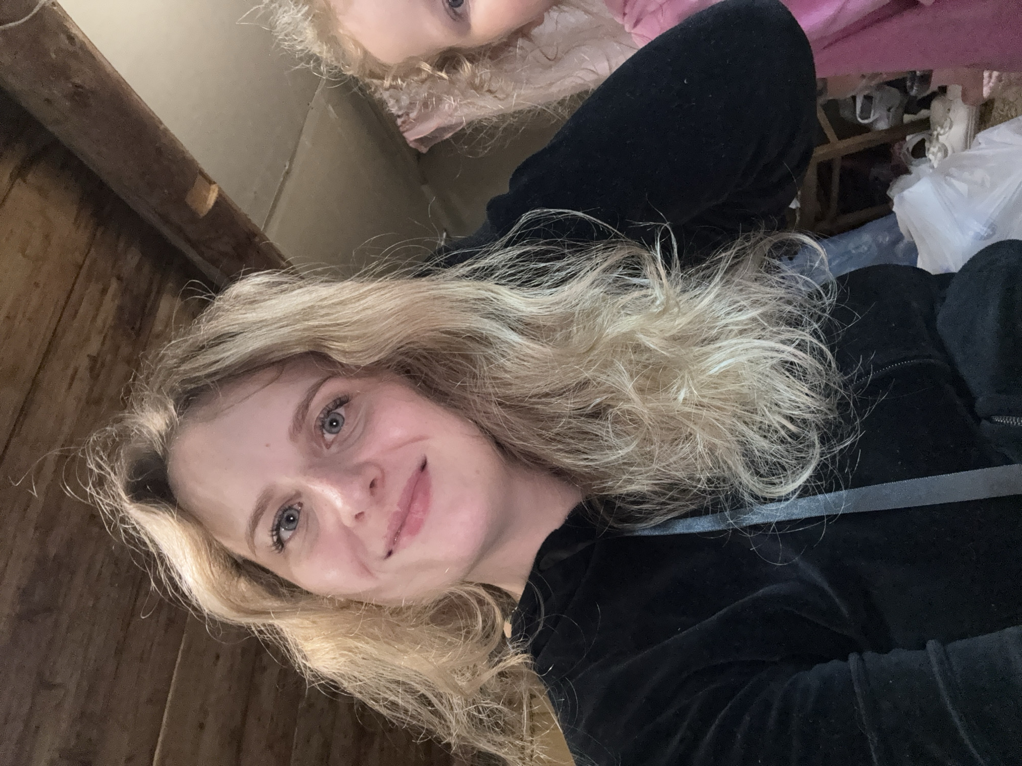 A medium close-up selfie shows a fair-skinned woman with blonde, wavy hair. She is wearing a black hooded jacket. To her right, a child with blonde curly hair is partially visible, wearing a pink garment. The background appears to be an indoor setting with a wooden ceiling and a cluttered floor with bags and shoes. The location is not specified in the image or metadata, and cannot be determined.