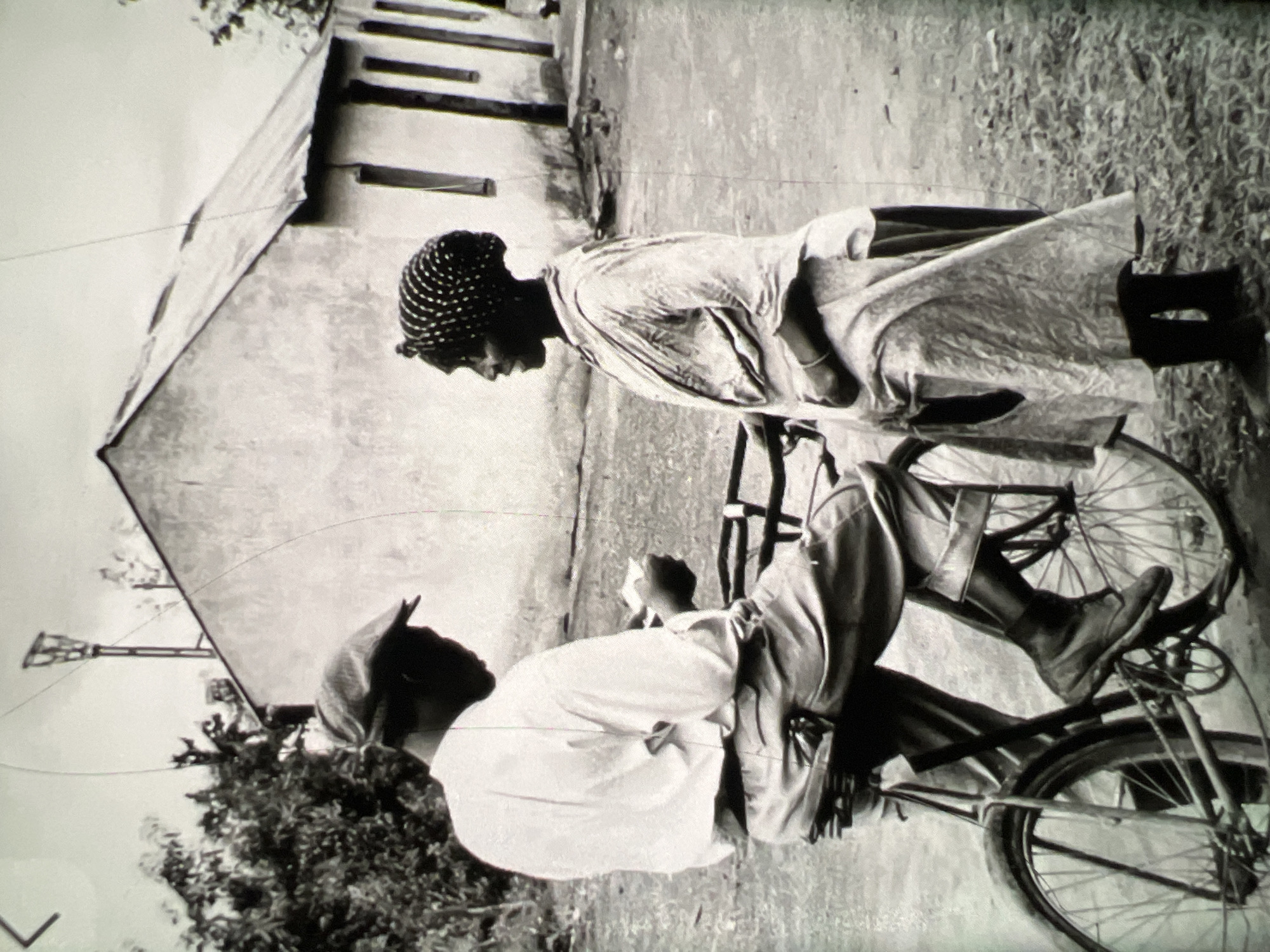 In this black and white photograph, a man wearing a flat cap and a light-colored shirt and trousers sits astride a bicycle. He is looking down at something in his hands. A woman, wearing a patterned headwrap and a patterned dress, stands facing him, looking in his direction. Behind them is a building with a corrugated metal roof and several windows. The scene appears to be outdoors, possibly in a rural or semi-urban setting in Lagos, Nigeria, based on the architectural style and vegetation. The ground is covered with dry grass and dirt. The overall impression is a candid moment captured between two individuals.