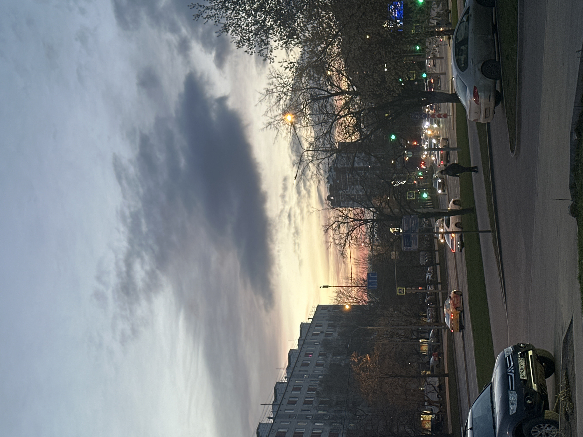 The image depicts a city street scene in Moscow, Russia at dusk. A large apartment building is visible along the right side of the frame. Trees with bare branches line the street. Cars are parked along the sidewalk and driving along the street. Street lights illuminate the area. Pedestrians are visible on the sidewalk. The sky is filled with clouds and the sunset is visible between the clouds and buildings.