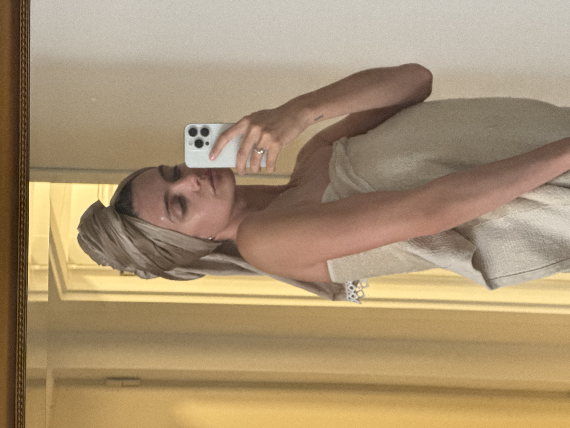 A mirror selfie featuring a woman wrapped in a towel. She also has a towel wrapped around her head. She is holding a white smartphone up to her face to take the picture. The background shows a pale yellow wall with a door or window frame. The mirror has a gold-colored frame.