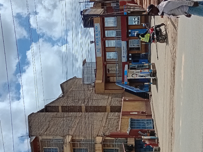 The image shows a street scene in Mlolongo, Kenya, located at coordinates -1.401 latitude and 36.944 longitude. It depicts a petrol station with a blue and white "DIESEL" pump on the left, and a building with a sign that reads "FILL & GO FUEL HERE" on the right. A man in a helmet is sitting on a motorcycle parked near the petrol station. A man in a white shirt and a blue cap is standing near the wood planks, facing the street. The environment consists of a paved road with a sidewalk, and buildings with exposed brickwork. The sky is partly cloudy with a few wispy clouds. Numerous power lines are strung across the scene.