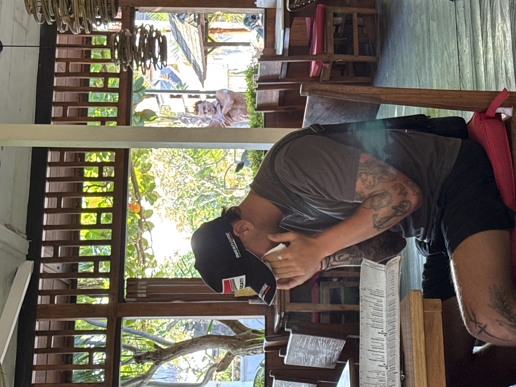 Inside a restaurant in North Kuta, Indonesia, a seated man in a grey t-shirt, black shorts, and a "Ducati Corse" baseball cap holds a tissue to his face while looking at a menu. His arms are tattooed. He is sitting at a wooden table with a red cushion. The restaurant has wooden latticework windows with views of lush greenery outside. Sunlight streams in, illuminating the space. A wooden chandelier hangs from the ceiling. Other tables and chairs are visible in the background. Beyond the windows, glimpses of a beach and sky are visible.