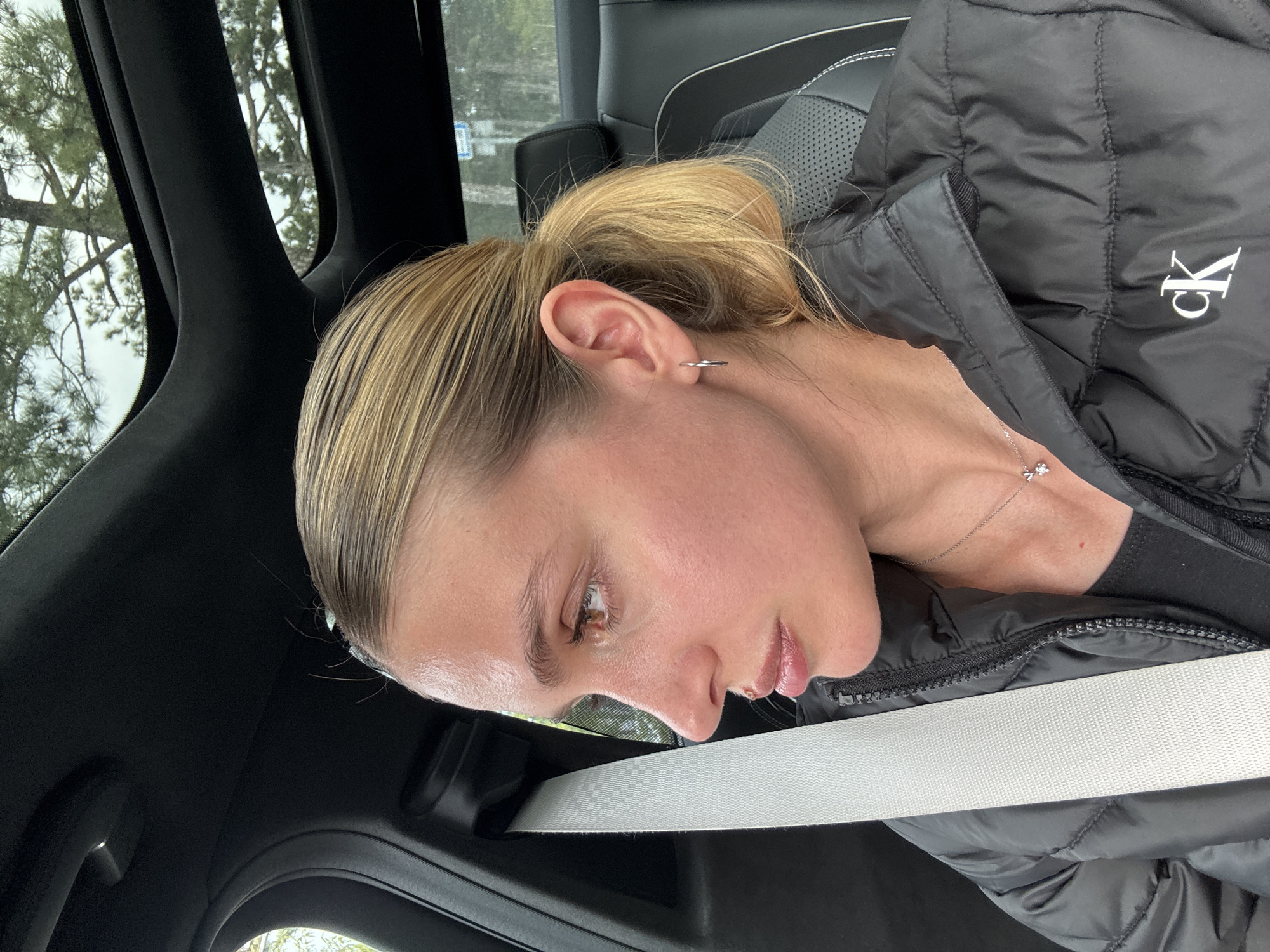 A close-up, rotated image shows a young woman sitting inside a vehicle, likely a car, with the specified location being Tbilisi, Georgia. Her blonde hair is pulled back in a ponytail, and she is seen in profile, looking downwards. She has a neutral expression, possibly contemplative.
She is wearing a black quilted jacket with a visible "ck" logo (representing Calvin Klein) on the upper left arm or shoulder area. Around her neck is a delicate silver necklace featuring a small, sparkling charm. A simple silver hoop earring adorns her left ear. A white seatbelt is securely fastened across her chest and lap.
The car's interior is visible, featuring dark, possibly leather, seats with perforated sections and dark trim on the pillars and ceiling. Through the window in the upper left, out-of-focus trees and an overcast sky suggest it is daytime with soft, diffused natural light. No specific activity beyond sitting in the car is discernible.