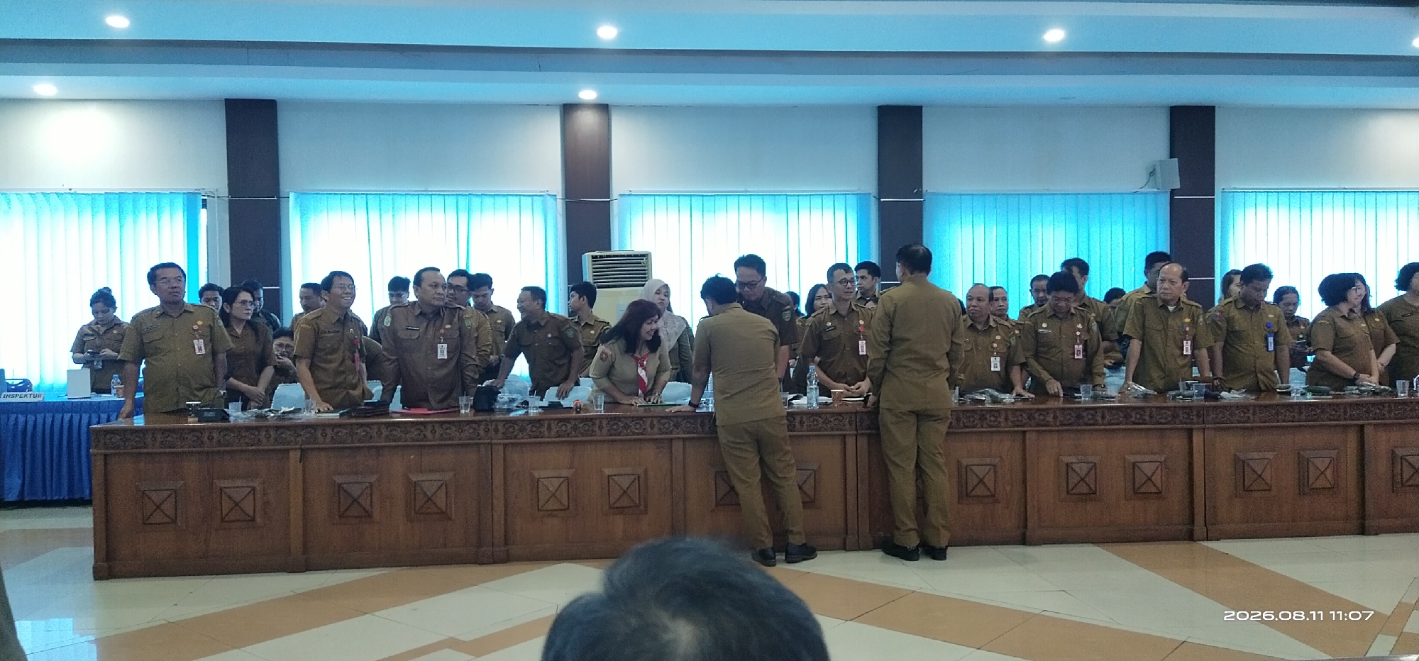 The image depicts a formal gathering of people, likely government officials or employees, in a conference room in Tamiang Layang, Indonesia. The individuals, mostly wearing matching brown uniforms, are standing behind a long wooden table. Many have ID cards or badges attached to their uniforms. The room is well-lit with overhead lighting, and the background features light blue curtains and a large air conditioning unit. The overall atmosphere suggests a meeting, presentation, or official event. The date and time stamp "2026.08.11 11:07" is visible in the bottom right corner.