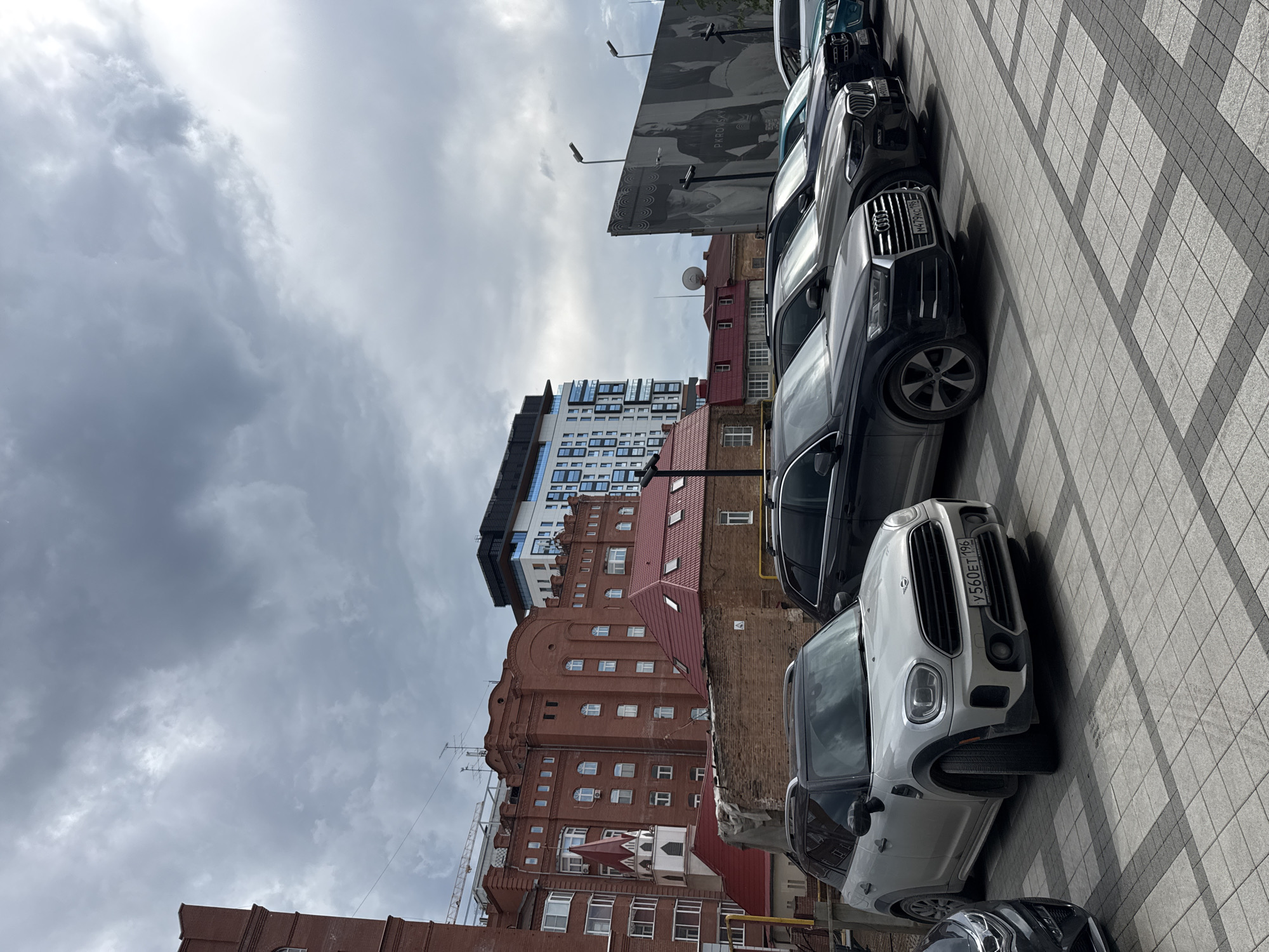 The image captures a row of parked cars in a city. The cars are of various makes and models, and they are all parked on a paved area. In the background, there are several buildings, including a tall, modern building and some older brick buildings. A large billboard with a black and white image of several people is also visible. The sky is cloudy, suggesting overcast weather. The license plate on the silver Mini Cooper reads "У560ЕТ 196". The license plate on the black Audi reads "M479KC 196". This scene is located in Yekaterinburg, Russia, likely in a commercial or residential area with a mix of new and old architecture.