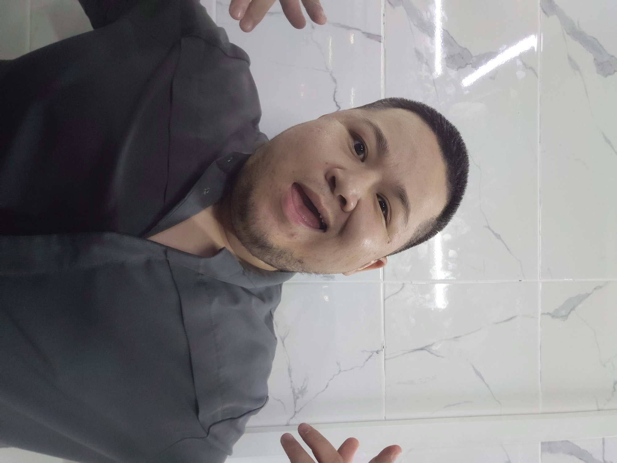 A medium shot shows a person with a shaved head, dark eyebrows, and an open mouth with their tongue visible, against a white tiled background with gray marble-like veining. The person is wearing a dark gray collared shirt with the top buttons undone. Their hands are raised with fingers splayed, palms facing outward, in a gesture that could be interpreted as playful or defensive. The lighting appears to be from above, casting bright reflections on the tiles. The image has a slightly distorted, wide-angle perspective, consistent with a selfie taken with a smartphone. The location is in the city of Aktau, Kazakhstan.