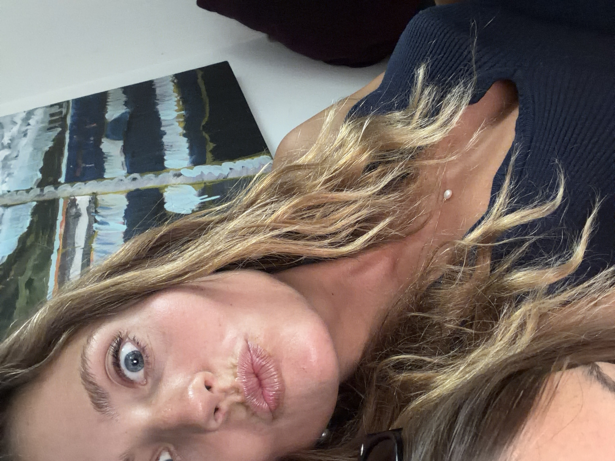 A selfie of a woman with blonde hair and blue eyes. She has her lips puckered in a kissing expression. She is wearing a dark blue top and a small pearl earring. An impressionist style painting of water is visible in the background. The location and time is uncertain.