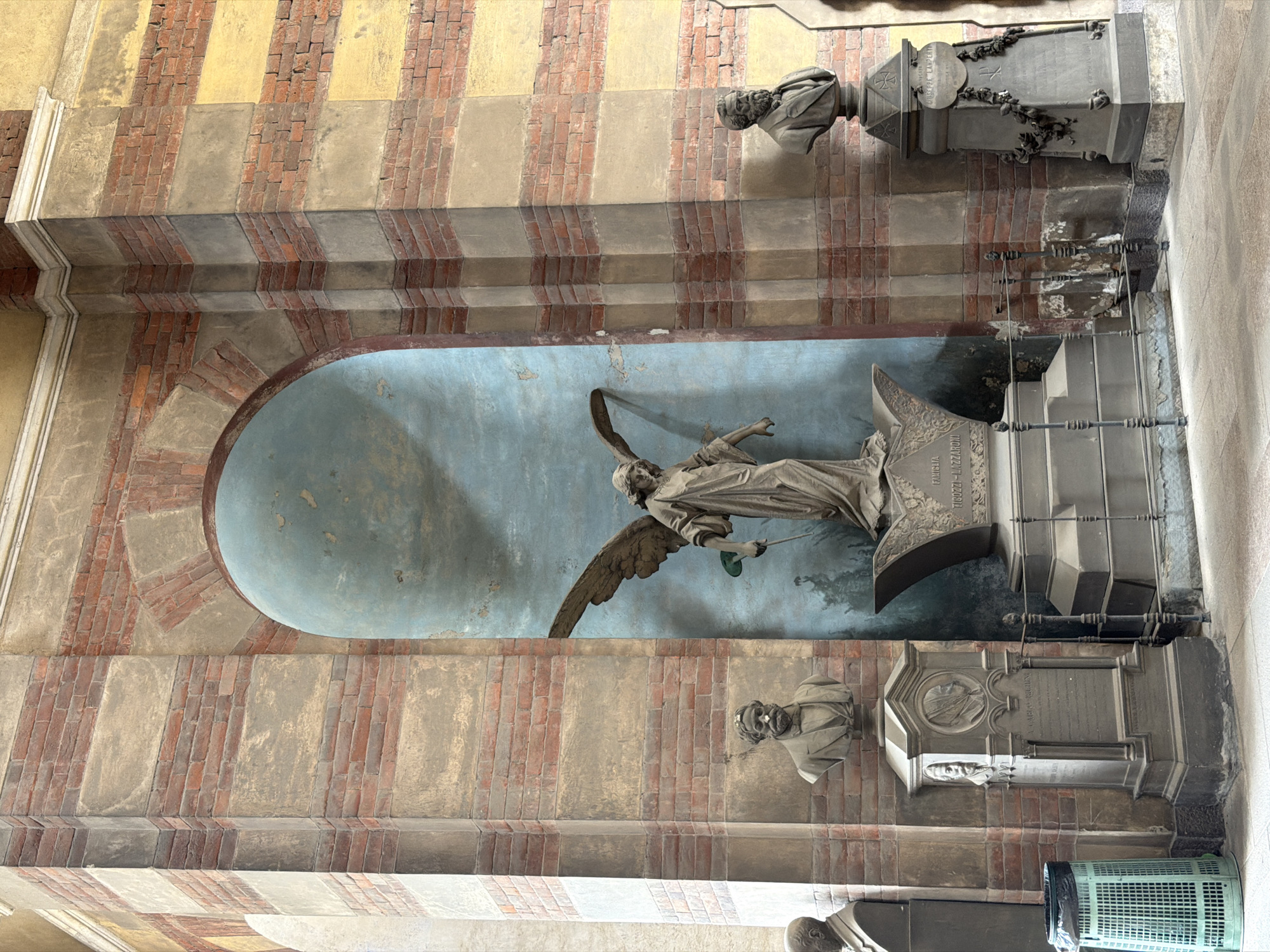 The image depicts a section of the Cimitero Monumentale in Milan, Italy. Visible are multiple burial monuments along a covered walkway. The architectural style of the walkway features striped walls of brick and stucco with arched niches.
The most prominent monument features a large marble sculpture of an angel with outstretched wings, set within a blue-painted arched niche. Below and above it are smaller monuments with sculpted busts and inscribed plaques. One tombstone has a metal railing fence.
The light is ambient and appears to come from the open side of the walkway, suggesting an outdoor environment with a roof or overhang. A green basket, possibly a trash can, is at the bottom right corner. The image is shot at eye-level, tilted to the left.