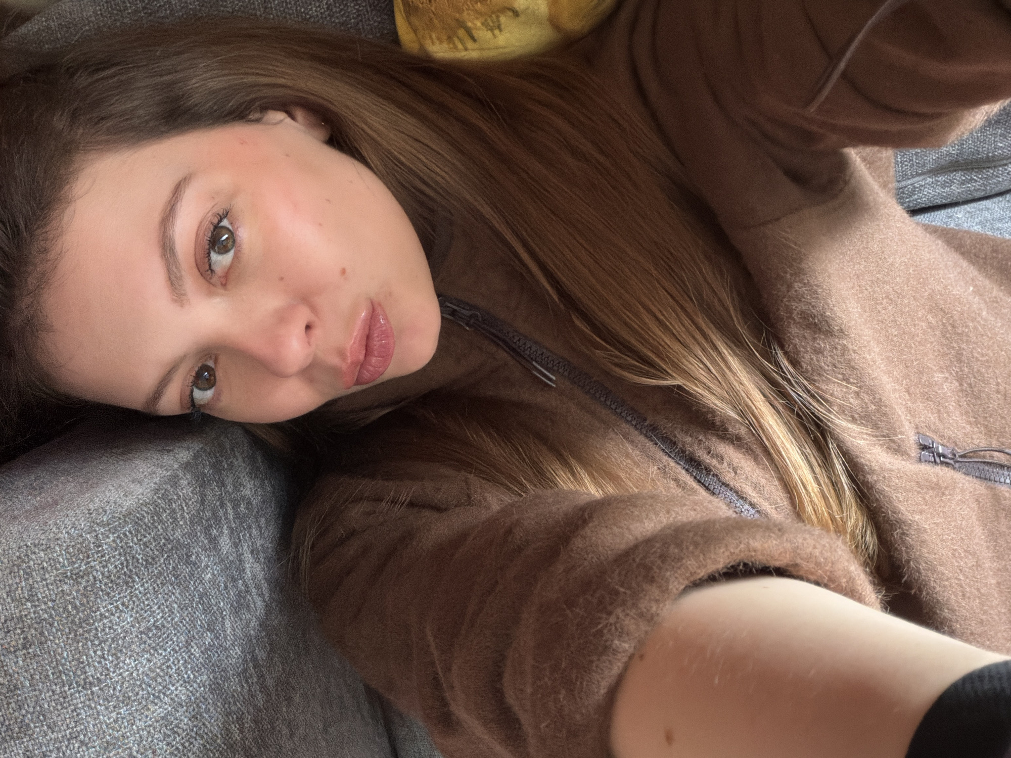 A young woman is captured in a close-up selfie, lying down in an indoor setting in Khimki, Russia. She is positioned diagonally on what appears to be a gray sofa or cushioned surface. The woman is wearing a textured, fuzzy brown zippered jacket or sweatshirt, with her long, light brown hair fanned out across her shoulders and clothing. Her gaze is directed towards the camera with a neutral to slightly contemplative expression, her lips subtly parted and a hint of blush on her cheeks. Light eye makeup and lip gloss are visible. In the upper background, a sliver of a yellow object, possibly a pillow or blanket, can be seen. The lighting in the image suggests it is daytime. There is no visible text.