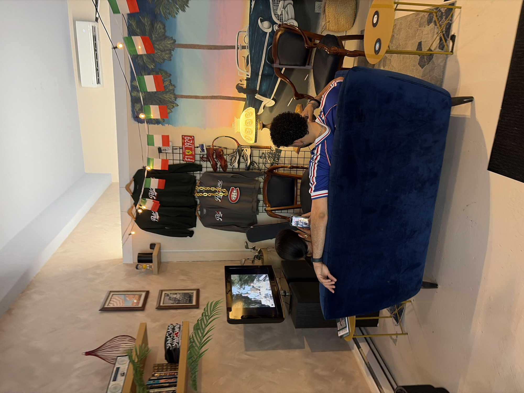 The scene depicts the interior of a room decorated with Italian-themed decor. A man with curly hair, wearing a blue and white jersey, is sitting on a dark blue velvet couch, looking at a phone held by a woman with dark hair next to him. A television mounted on the wall displays an image of a scene, possibly a movie or show. The room features vintage-style furniture and accessories, including a small, round, yellow side table, wooden chairs, and clothing items hanging on a rack. A mural depicting a coastal scene with palm trees and Italian flags adorns the wall. Other decor includes framed pictures, a bookshelf with various objects, and an air conditioning unit on the upper wall. The location is likely in Montreal, Canada, suggested by the Montreal Canadiens apparel on the rack.