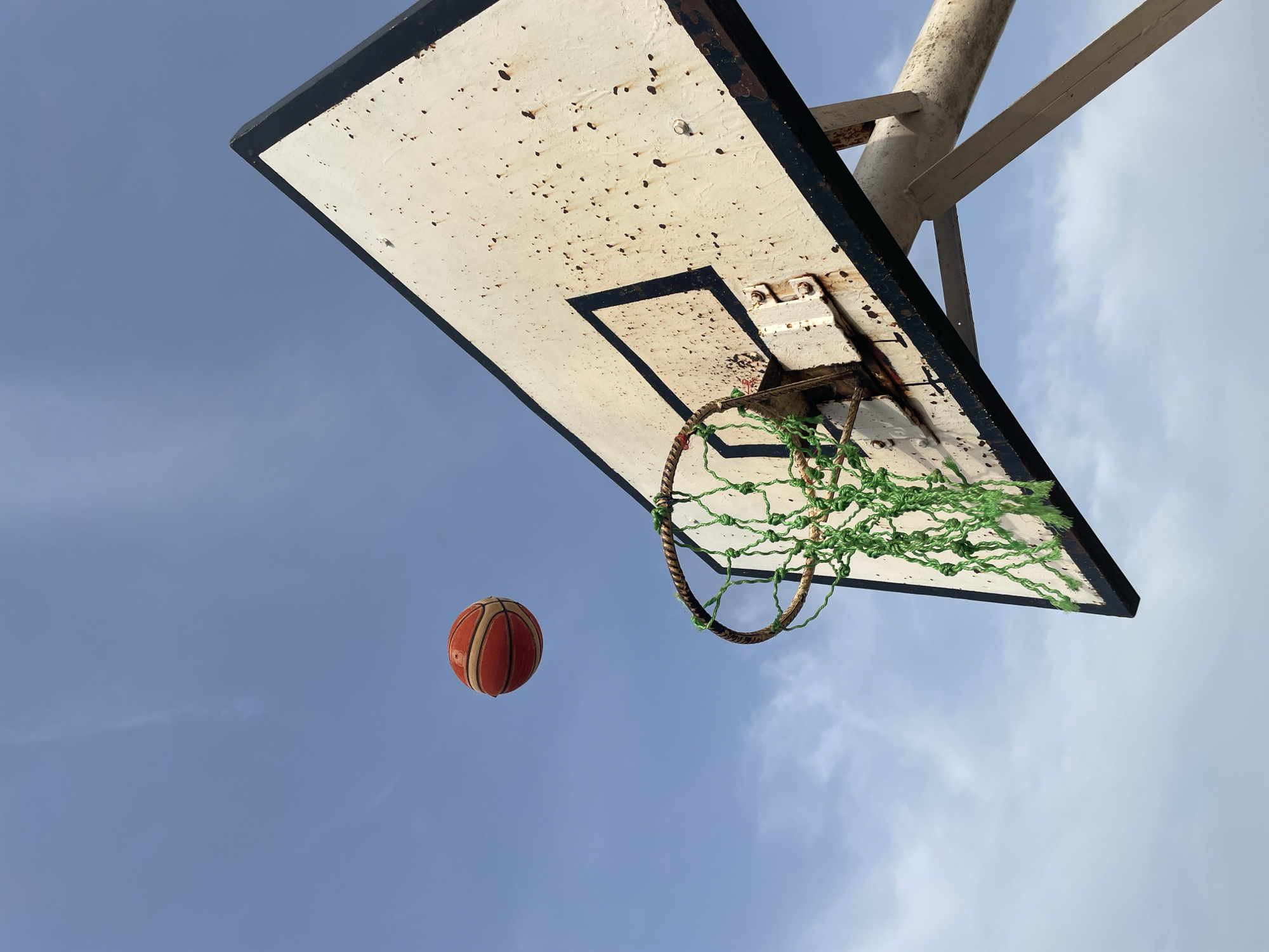 A low-angle shot shows a rusty basketball hoop against a blue sky. The backboard is white with a black border and rust spots. The hoop has a green net made of rope. An orange basketball is suspended in the air below the hoop. The location is Akwanga, Nigeria.