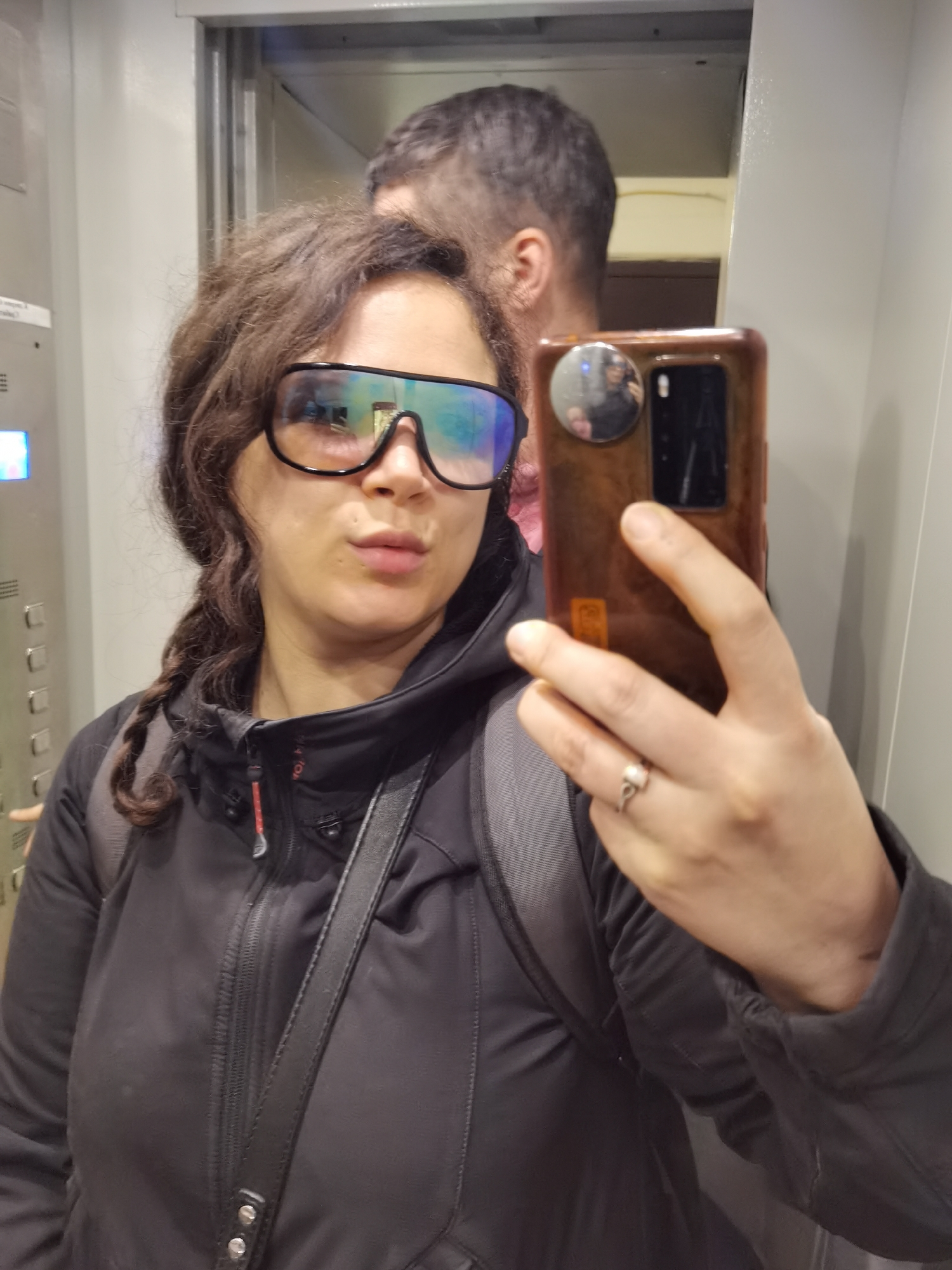 A woman with curly brown hair, partially styled into a braid, is capturing a mirror selfie in what appears to be an elevator. She is wearing large, mirrored, rectangular sunglasses with a black frame and a black zippered jacket over a dark backpack. Her lips are slightly puckered, suggesting a playful or casual expression. In her right hand, she holds a brown-cased smartphone, which features a circular pop socket or holder and an indistinct sticker or QR code on the back. A silver-colored ring with a light-colored, possibly pearl or opal, stone is visible on her right ring finger.
Behind the woman, the reflection of a man is visible in the mirror. Only the back of his head, showing short dark hair and a beard, is seen. His reflection is also faintly discernible in the woman's mirrored sunglasses.
To the left, an elevator control panel with a silver-grey surface features multiple square buttons arranged vertically. A small digital display on the panel emits a bright blue or purple light, but no specific numbers or text are legible. The reflective walls of the elevator suggest a metallic or polished surface.
The scene takes place indoors, within an elevator. The woman's pose and expression convey a lighthearted and casual mood. No specific time of day or weather cues are evident. The image contains no legible text. The provided location context is Chaikovsky, Russia, but no visual elements in the image confirm this geographical detail.