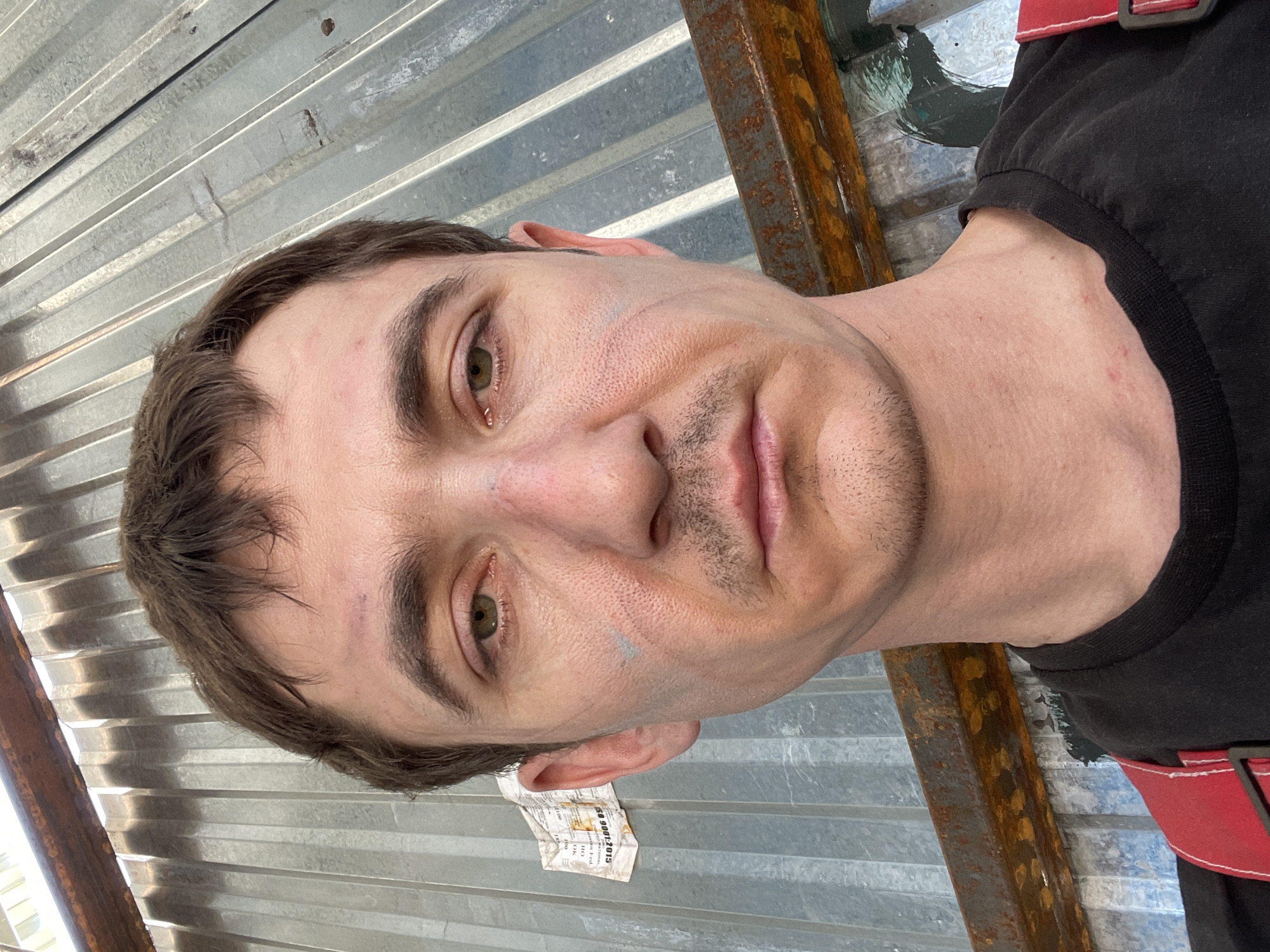 The image is a close-up, selfie-style photograph of a man's face and upper chest. He has short, brown hair and appears to be outdoors, with a corrugated metal wall as the background. There are some smudges of what looks like dirt or paint on his face, specifically around his eyes and cheeks, and a small bruise on his forehead. He is wearing a black t-shirt with red suspenders. The lighting suggests it is daytime. No specific location within the city of Сокуры, Russia can be determined from the image.