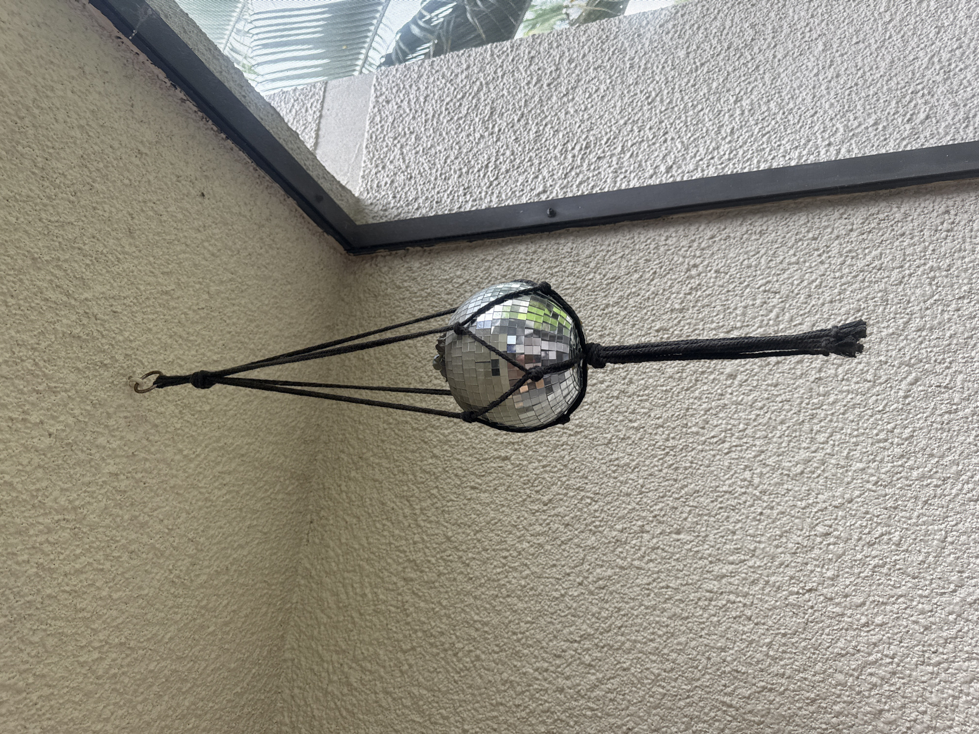 A disco ball, suspended from a black macrame hanger, hangs in front of a textured, light-colored wall. The disco ball is composed of numerous small, reflective square mirrors. The hanger is attached to a ceiling hook. To the right of the disco ball, a dark-colored window frame is visible, with a screen and the reflection of greenery beyond it. The setting appears to be an outdoor or semi-outdoor space, possibly a balcony or porch, given the wall texture and the presence of a window and screen. No people or animals are present in the image. The scene is static and shows a decorative object.