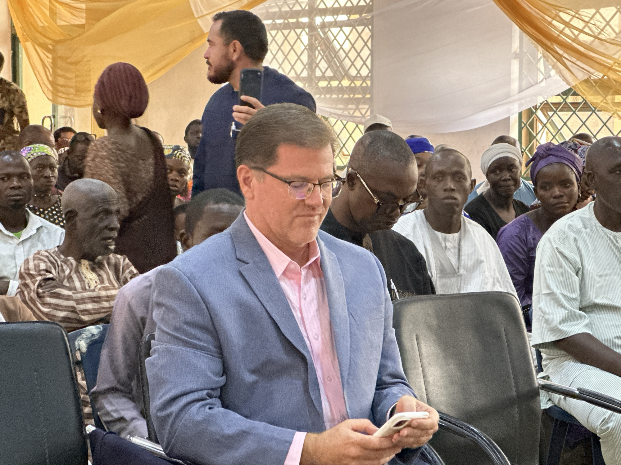 A photo taken in Unknown, Unknown, shows a crowd of people indoors. The main subject is a man in the foreground, wearing a light blue suit jacket over a pink shirt and glasses, who is looking down at a smartphone he is holding. To his right, another man is wearing dark sunglasses and a black shirt. A third man stands in the background, holding a smartphone up to his face, wearing a dark blue top. The crowd of people are of different ages and dressed in various traditional and contemporary clothing. Overhead, a sheer yellow drape is suspended near a window with a grid pattern. The lighting is bright. The crowd are seated in chairs.