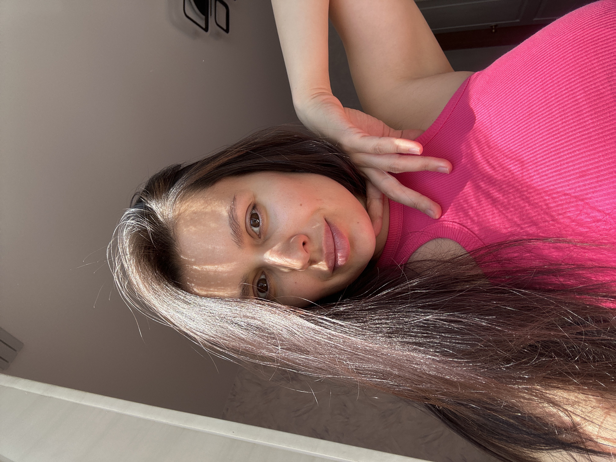 A young woman with long brown hair and light skin is looking directly at the camera. She is wearing a pink ribbed top with a small cutout at the shoulder. Her right hand is raised and positioned near her face, with her fingers slightly bent. Light streams across her face, creating shadows and highlights. The background consists of a white wall with a dark geometric shape visible in the upper left corner and a white ledge along the bottom edge of the image. There is no indication that the photo was taken in the city of Chaikovsky, Russia.