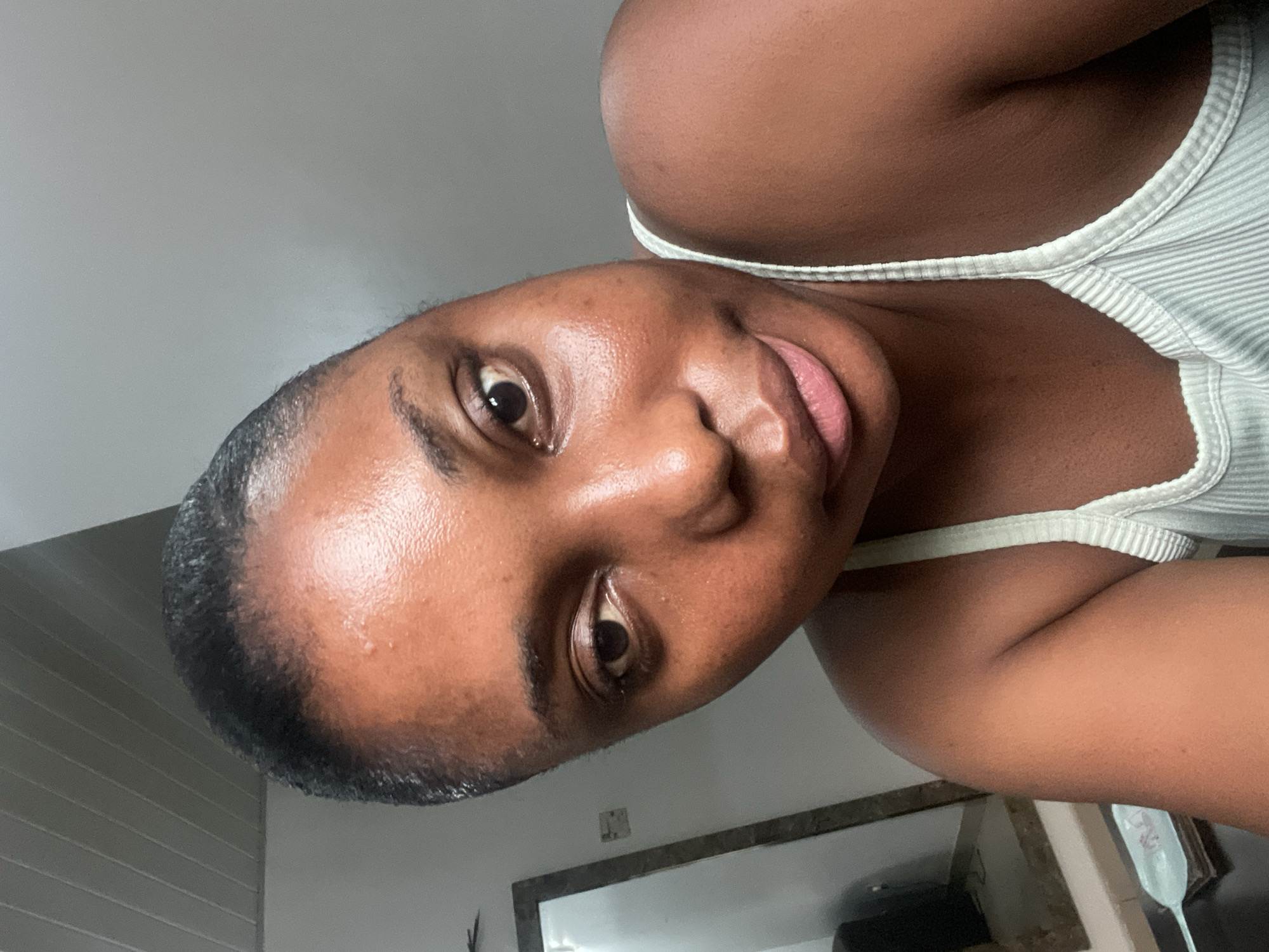 A medium close-up portrait shows a Black woman. The subject is fair complexioned, and is wearing a light-colored ribbed tank top. She is looking directly at the camera with a slight smile. The background is out of focus but suggests an interior setting. A portion of a mirror frame is visible in the lower part of the frame. The location, city of Abuja, Nigeria, is not deducible from the visual information.