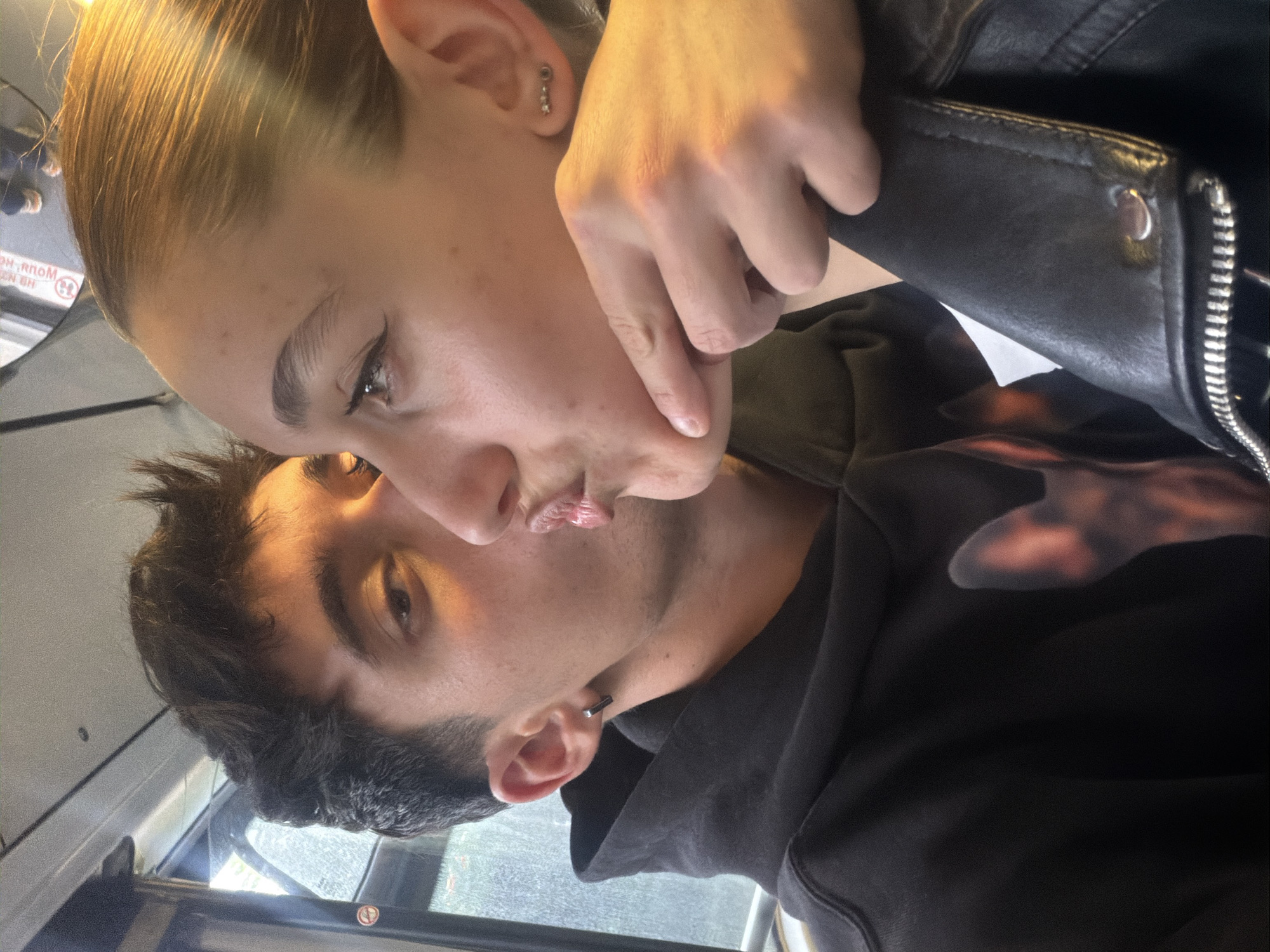 A close-up, rotated selfie shows two people. The person on the left has dark hair and is wearing a black hoodie with a blurred pattern. The person on the right has light-colored hair pulled back and is wearing a black leather jacket. The hand of the person on the right is pinching the cheek of the person on the left, forming a kissing expression. Both have pierced ears. The background shows a window and part of a bus or tram interior. A sticker with Cyrillic text and a public transport symbol is visible. It is not possible to confirm the precise location in Sofia, Bulgaria, based only on the provided image.