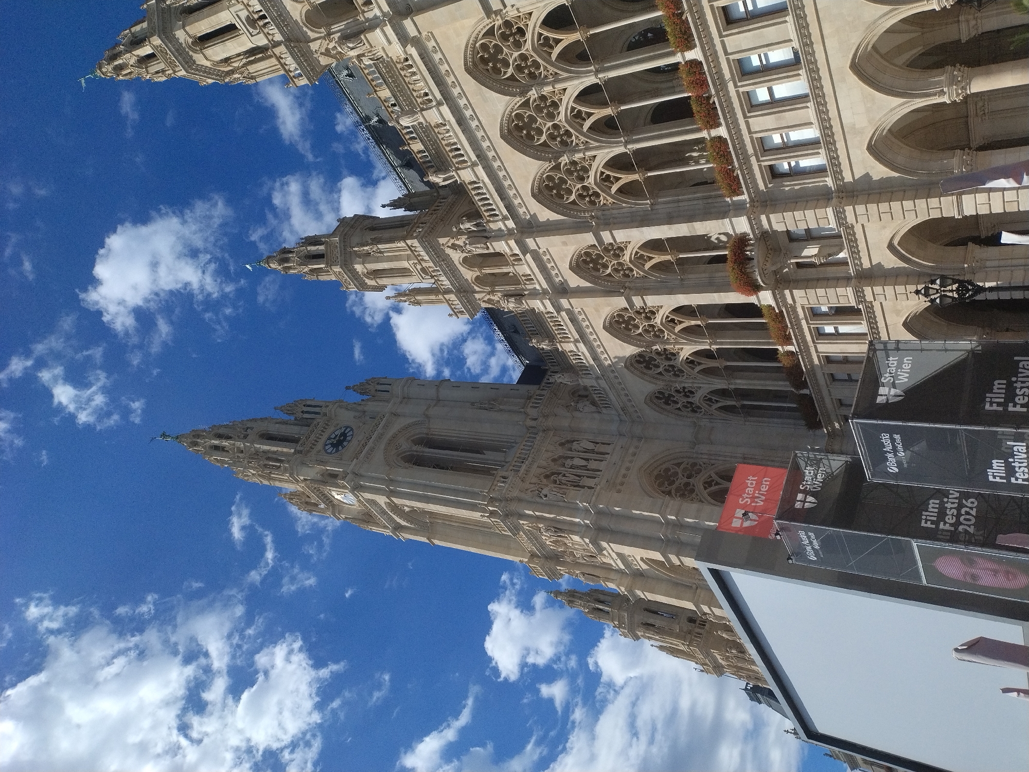 The image captures a low-angle view of the Rathaus (City Hall) in Vienna, Austria, under a bright blue sky with scattered white clouds. The ornate Gothic architecture of the Rathaus dominates the frame, with its intricate stonework, pointed arches, and multiple spires. A prominent clock tower is visible on the left side of the building.
In the foreground, a large, blank white screen is set up, suggesting an outdoor event, possibly a film festival. Next to the screen, several banners and scaffolding are present. One banner prominently displays "Stadt Wien" (City of Vienna) in red and white, along with "Bank Austria UniCredit" and "Film Festival 2026." A partial image of a person's face is visible on one of the banners. The overall scene suggests a civic building hosting a public event.
