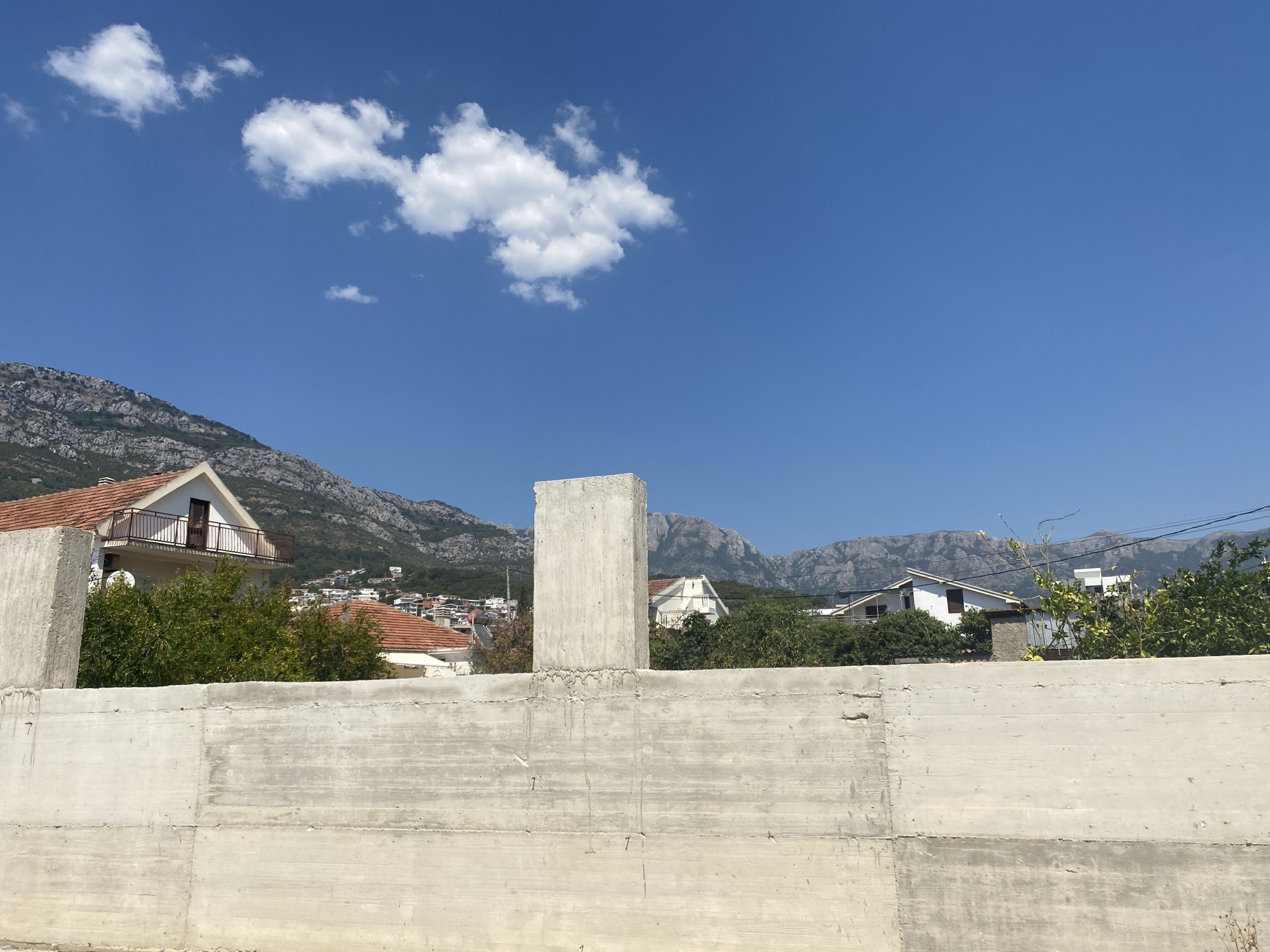 The image shows a bright, sunny day with a clear blue sky dotted with a few white clouds. In the foreground, there is a rough, unfinished concrete wall, likely part of a construction site. To the left of the wall, a tall, rectangular concrete pillar stands erect. Behind the wall, several houses with orange-tiled roofs are visible, nestled amongst lush green trees. In the background, a mountain range with rocky slopes forms a dramatic backdrop, suggesting a location near a mountainous coastline. The architecture of the houses and the overall landscape are consistent with the city of Bar in Montenegro, a region known for its coastal mountains and Mediterranean-style buildings. There are no people or animals visible in the scene. The image appears to be a still photograph captured outdoors.