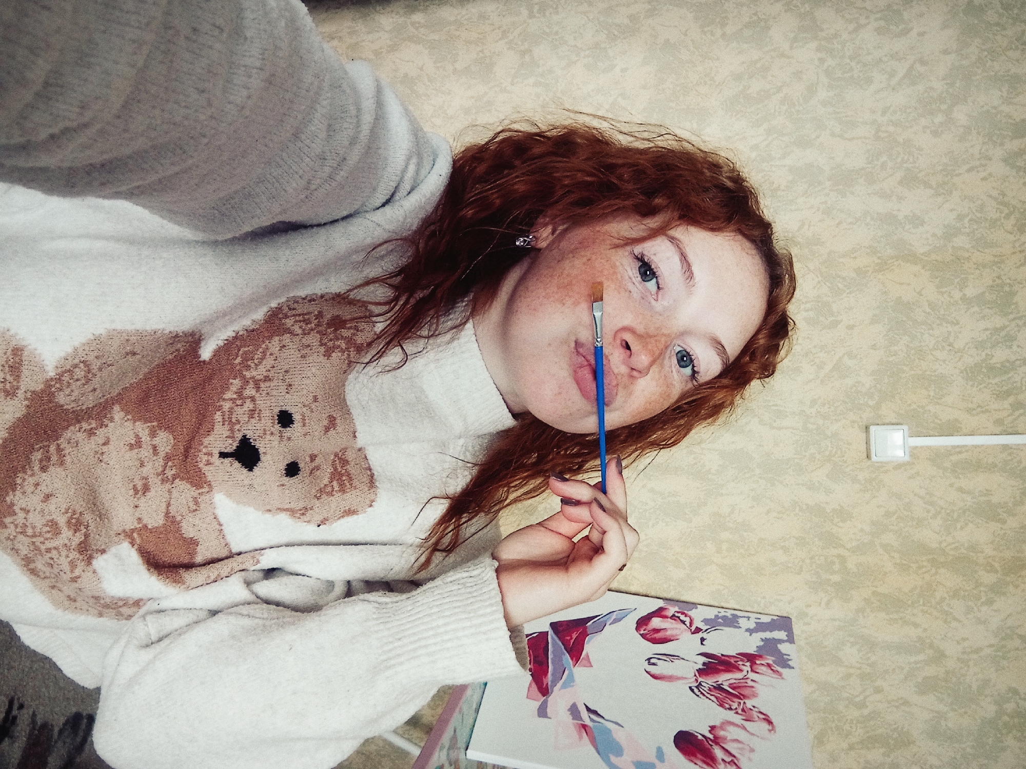 An overhead shot captures a young woman with reddish-brown, wavy hair, fair skin, freckles, and light blue eyes, looking upwards towards the camera. She is wearing a light-colored, possibly white or light grey, turtleneck sweater featuring a prominent brown and beige teddy bear design.
She holds a blue-handled paintbrush between her lips and chin, appearing pensive or thoughtful. To her lower right, an art canvas or board displays a floral pattern, possibly tulips, rendered in shades of pink, red, purple, and blue. The background is a textured, light-colored surface, suggesting an indoor setting like a wall or carpet. In the upper right corner, a white rectangular object, possibly a light switch or electrical outlet, is visible with a white wire extending from it. No text is visible in the image. The scene conveys a moment of quiet artistic contemplation in a domestic environment.