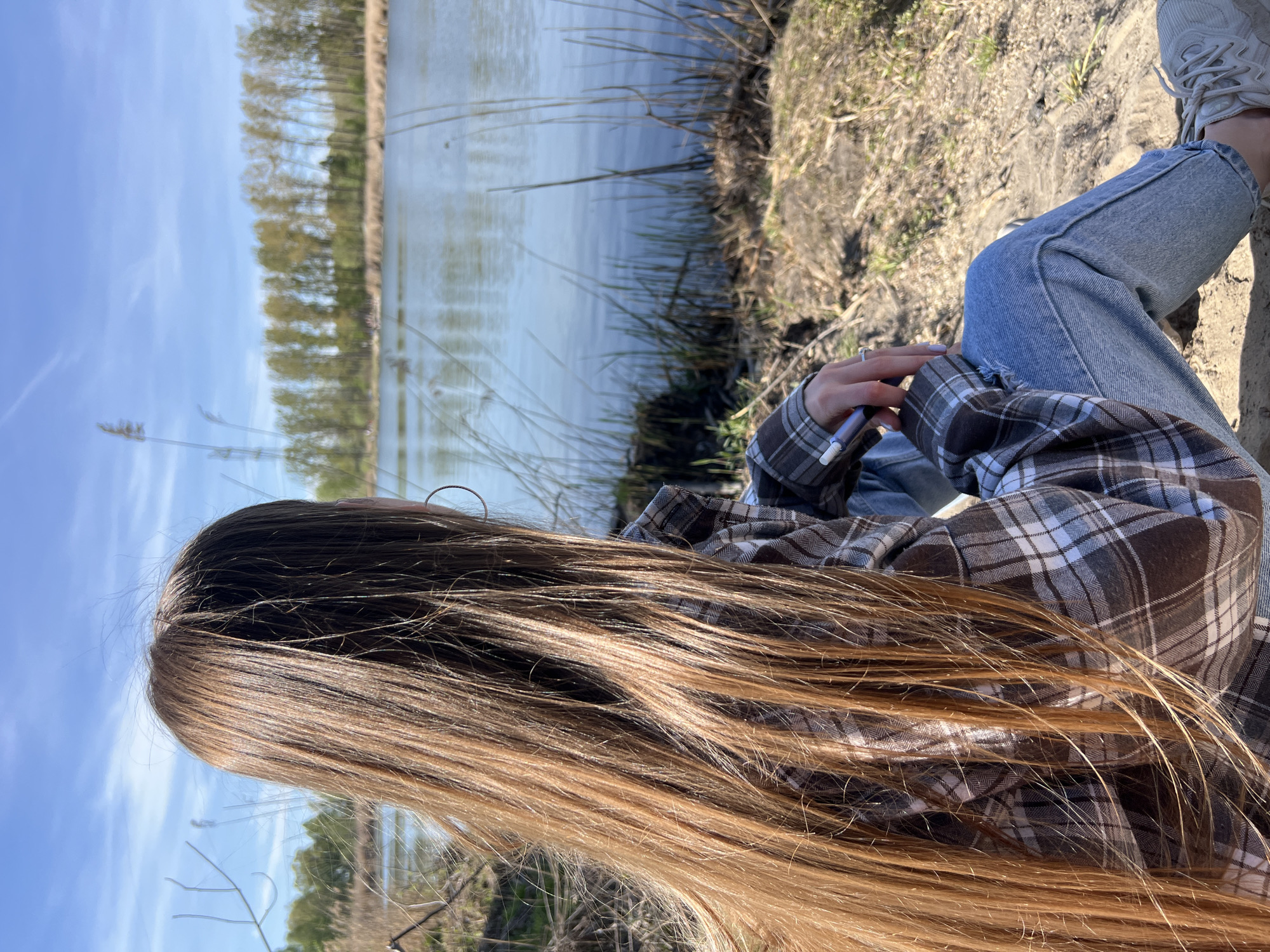 A person sits by a body of water on a sunny day. The person, seen from the side and back, has long brown hair, is wearing blue jeans, a brown and white plaid shirt, and white sneakers. They are holding a blue vape pen in their hands. The body of water is likely a lake or river, with trees lining the opposite bank. The ground around the person is sandy and has some vegetation. The sky is blue with some clouds. The location is likely in Ciemne, Poland.