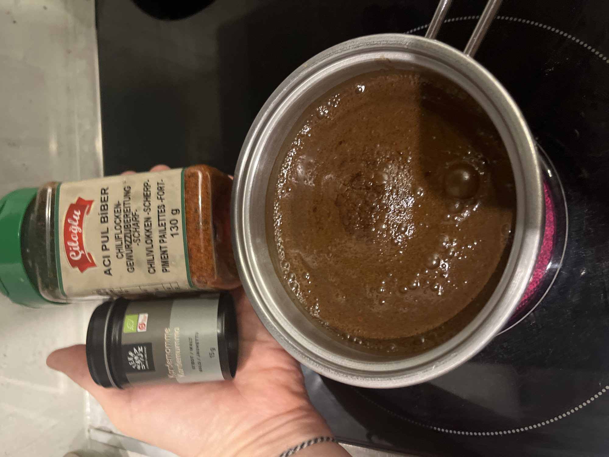 In a kitchen setting, a person's hand holds two spice jars next to a small metal pot on a black induction or electric stovetop. The pot contains a dark, frothy liquid, possibly coffee or a spiced drink, which appears to be heating, indicated by a faint reddish glow around the burner.
The larger spice jar, held by the fingertips, contains red pepper flakes. Its label, partially visible, reads "Ciloğlu ACI PUL BİBER" (Hot Flaked Pepper) with further descriptions in German ("CHILIFLOCKEN-GEWÜRZZUBEREITUNG -SCHARF-"), Dutch ("CHILIVLOKKEN-SCHERP-"), and French ("PIMENT PAILLETTES-FORT-"). The net weight is stated as "130 g".
The smaller, cylindrical spice jar, held firmly in the palm, is grey and black, and contains ground cardamom. Its label clearly shows "Kardemomme" (cardamom in Norwegian/Danish) and "Kardemumma" (cardamom in Swedish), along with "15g" indicating its weight. A visible organic certification logo is present on this jar.
The activity taking place appears to be the preparation of a beverage or food item, with spices being selected, likely to be added to the liquid in the pot. The image captures a moment of culinary preparation. The scene is set indoors, without specific time-of-day indicators beyond artificial lighting. The location context for this activity is Budva, Montenegro.